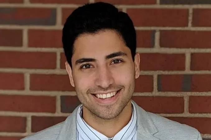 A young man with dark hair smiling in front of a brick wall, wearing a light gray blazer and a striped shirt.