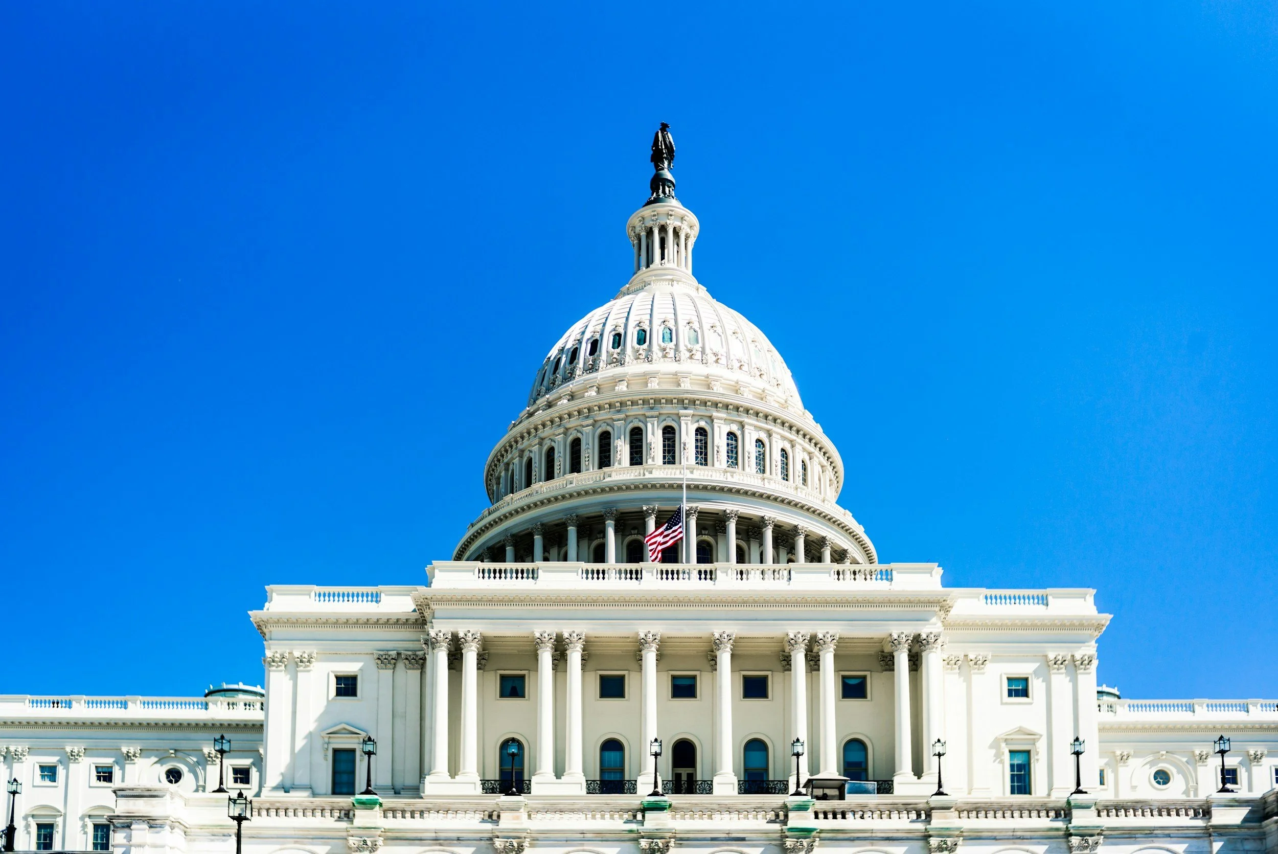 The United States Capitol building with a blue sky background and an American flag in front.