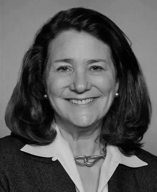 Black and white portrait of a middle-aged woman with shoulder-length hair, smiling, wearing a suit jacket, a collared shirt, a chain necklace, and pearl earrings.