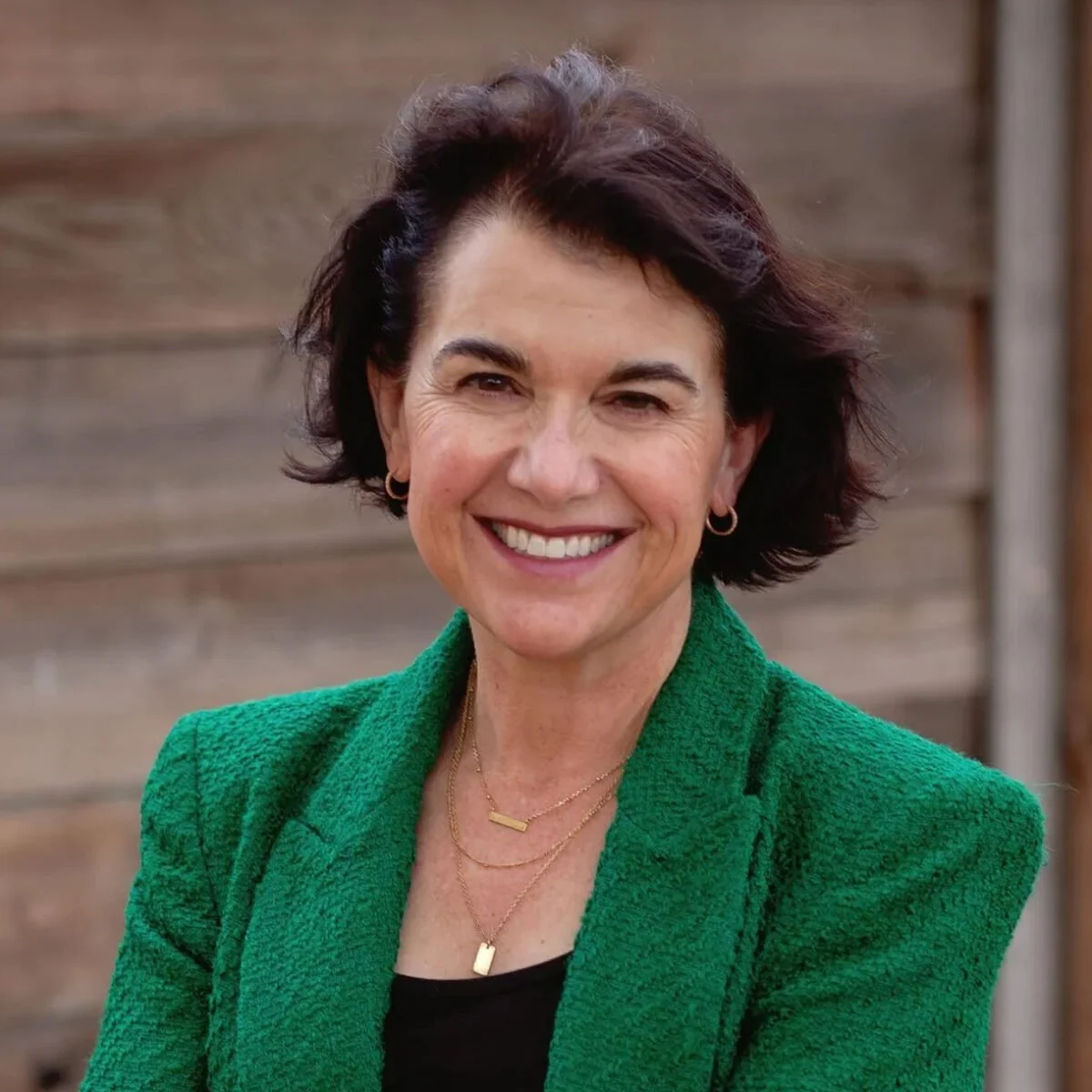 A woman smiling, wearing a green blazer, gold jewelry, and standing outdoors with a wooden background.