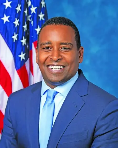A smiling man in a blue suit and tie, standing in front of American flags and a blue background.
