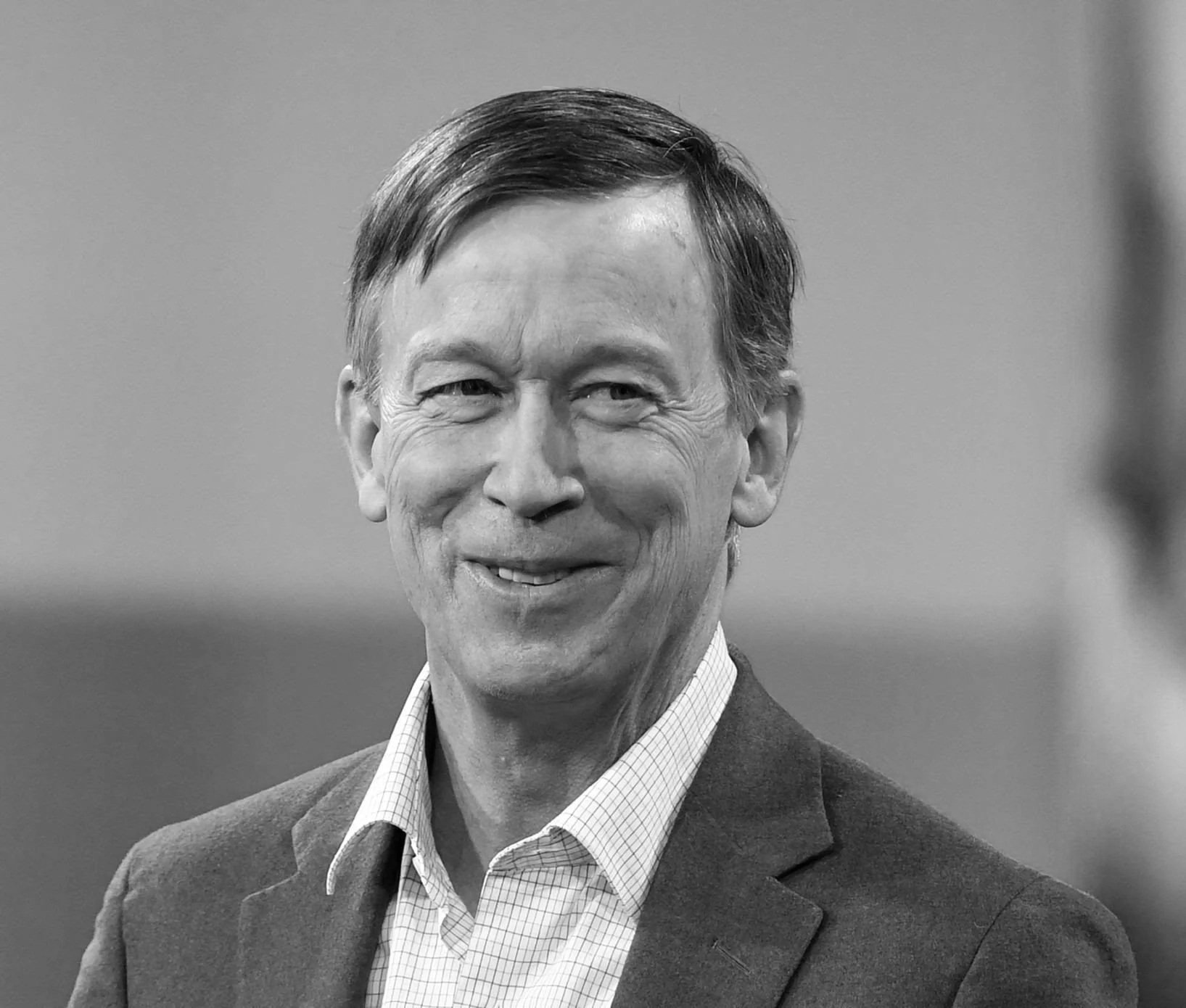 A black and white portrait of a smiling middle-aged man with short dark hair, wearing a suit and checkered shirt.