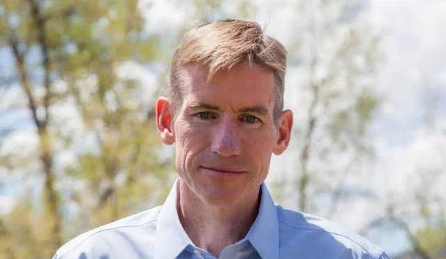 A man with light brown hair slicked to the side, wearing a light blue collared shirt, standing outdoors with trees in the background on a sunny day.