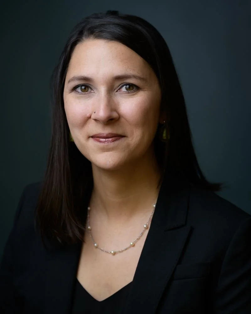 Professional portrait of a woman with dark hair, wearing a black blazer and jewelry, against a dark background.