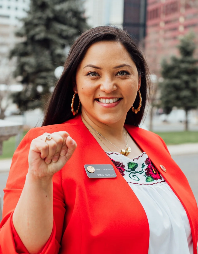 A woman wearing a red blazer and a name tag that reads 'Lupe C. Gonzales, State Senator' smiling and making a fist gesture outdoors with trees and buildings in the background.