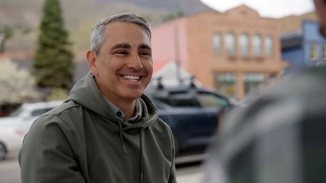 A smiling man with gray hair wearing a green jacket outdoors, with cars and colorful buildings in the background.