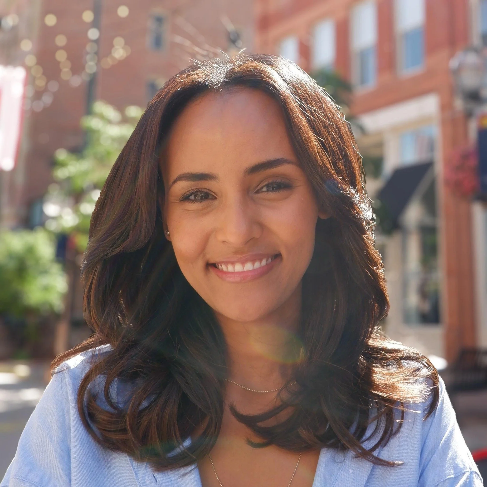Close-up of a smiling woman with wavy brown hair, wearing a light blue top, outdoors with blurred buildings and string lights in the background.