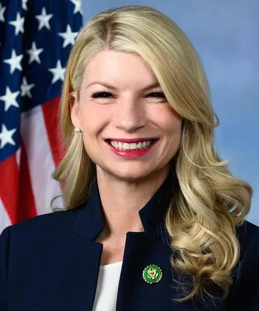 A smiling blonde woman in a navy blazer with a U.S. flag pin, standing in front of a waving American flag.