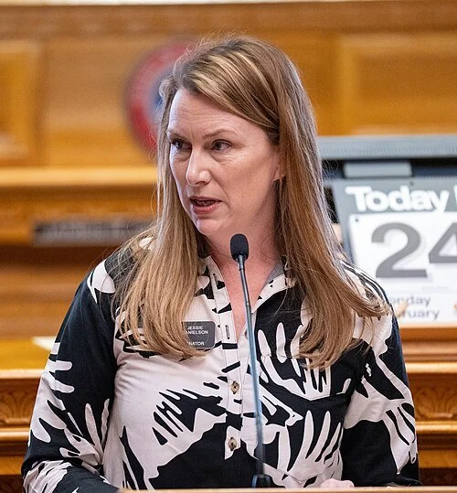Woman speaking into a microphone during a public event or hearing, with a wooden wall and a calendar in the background.