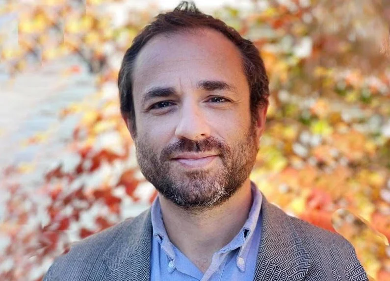 A man with a beard and brown hair smiling outdoors with autumn-colored leaves in the background.