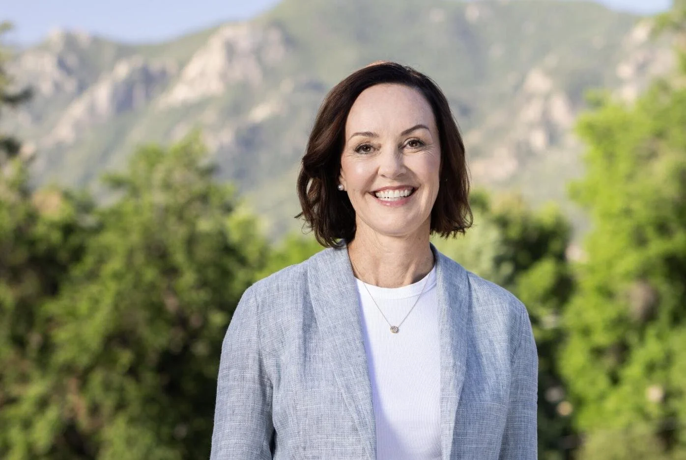 A woman with shoulder-length dark hair smiling outdoors, with mountains and green foliage in the background.