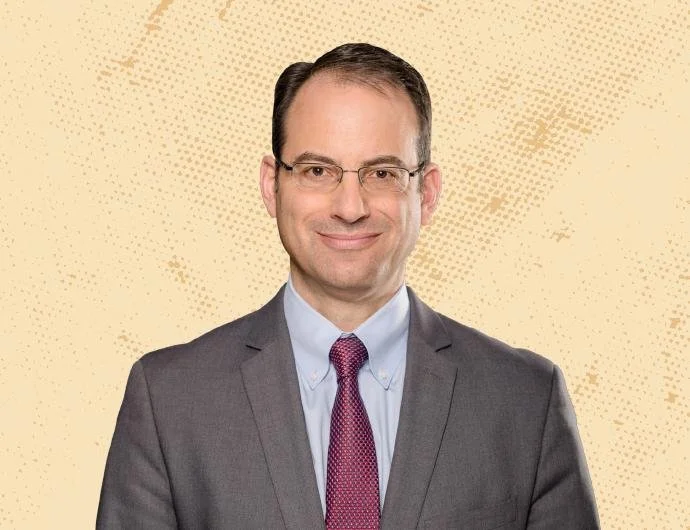 A man in a dark grey suit, light blue shirt, and purple tie, smiling at the camera, with short dark hair and glasses, against a beige dotted background.