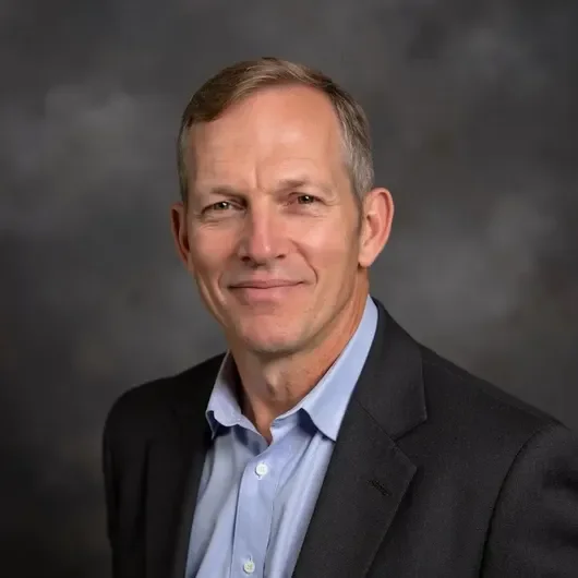 A middle-aged man with short brown hair, wearing a dark blazer and light blue collared shirt, smiling at the camera with a gray background.