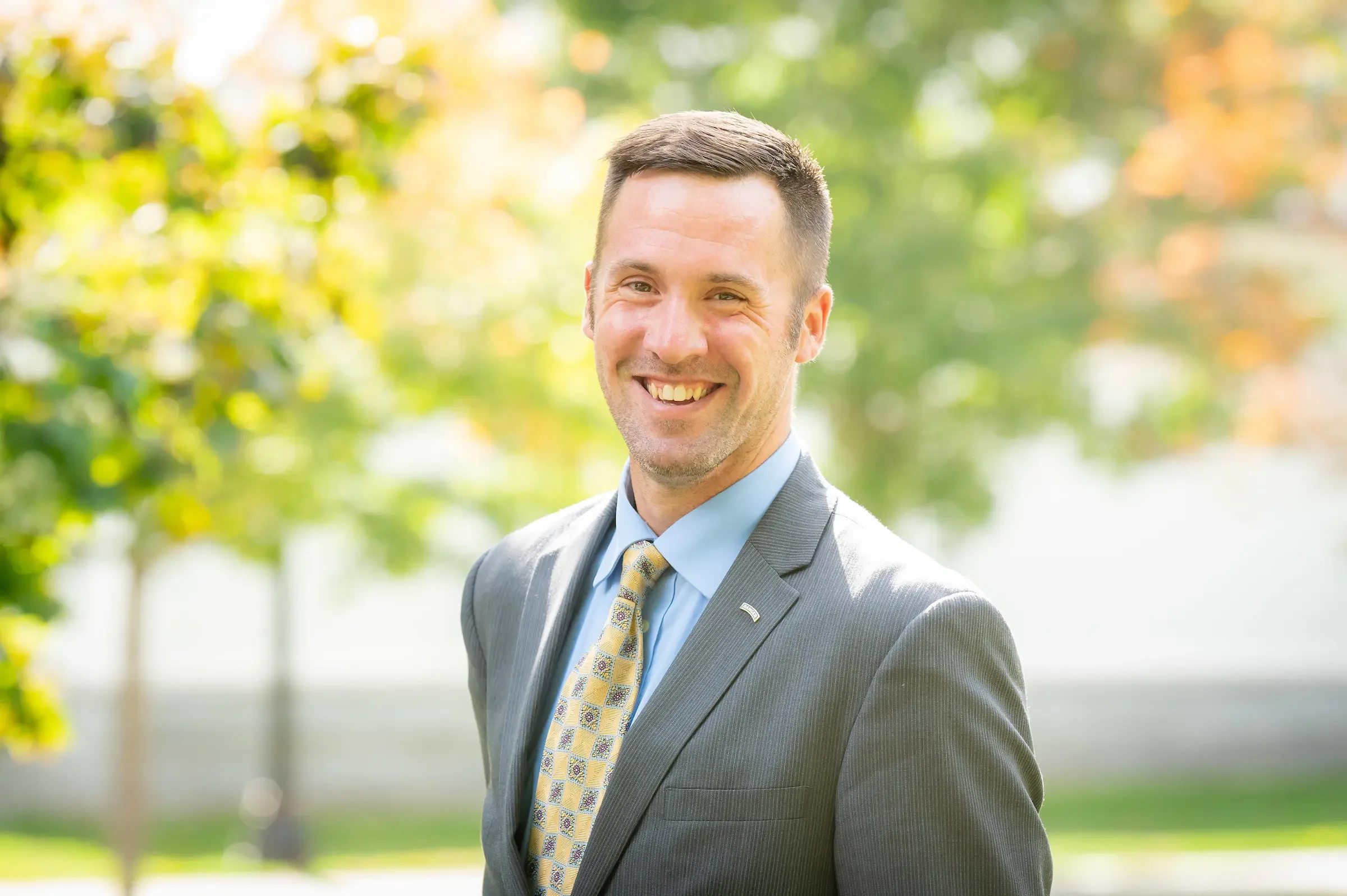A smiling man wearing a suit and tie, outdoors with blurred green trees and sunlight in the background.