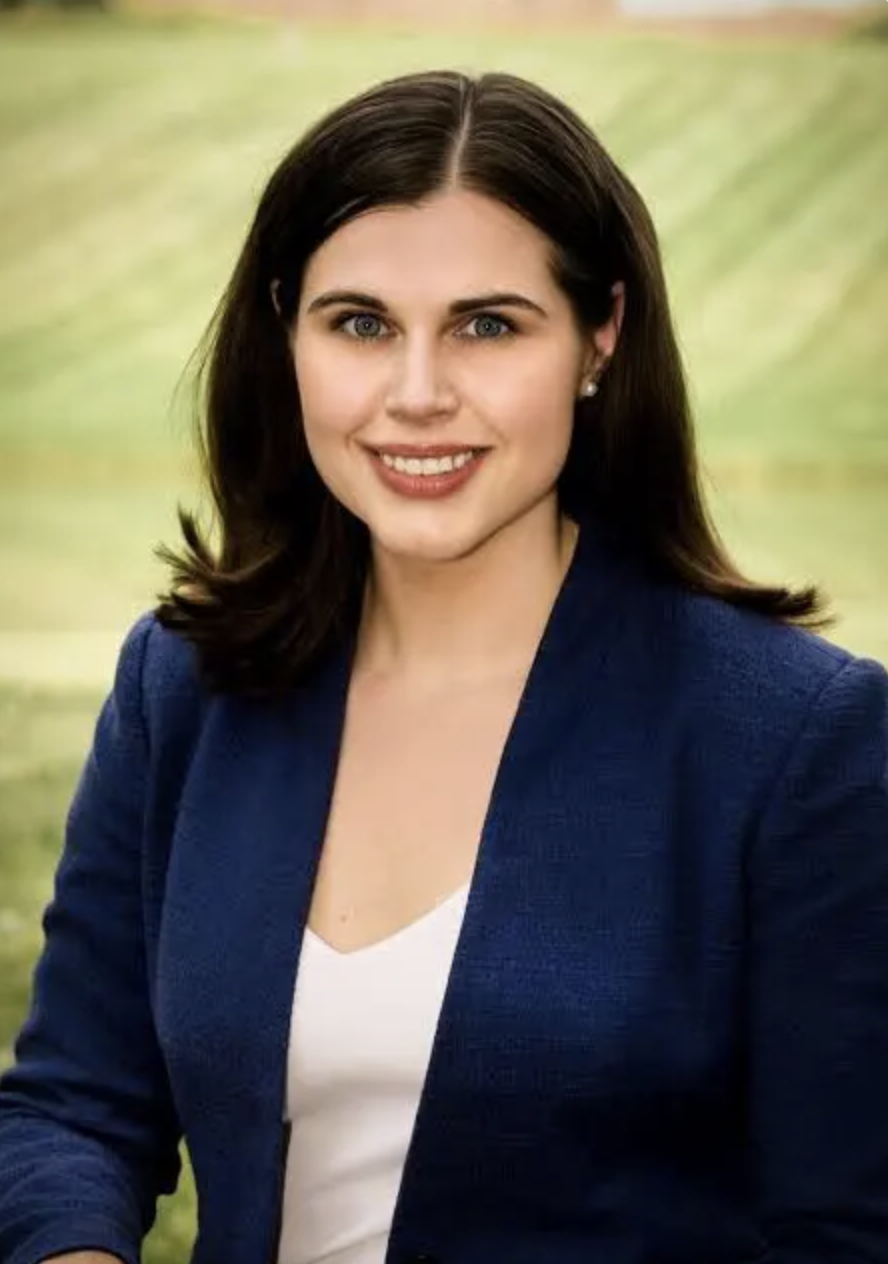 A woman with dark brown hair, wearing a navy blue blazer and a white top, smiling with a scenic outdoor background.
