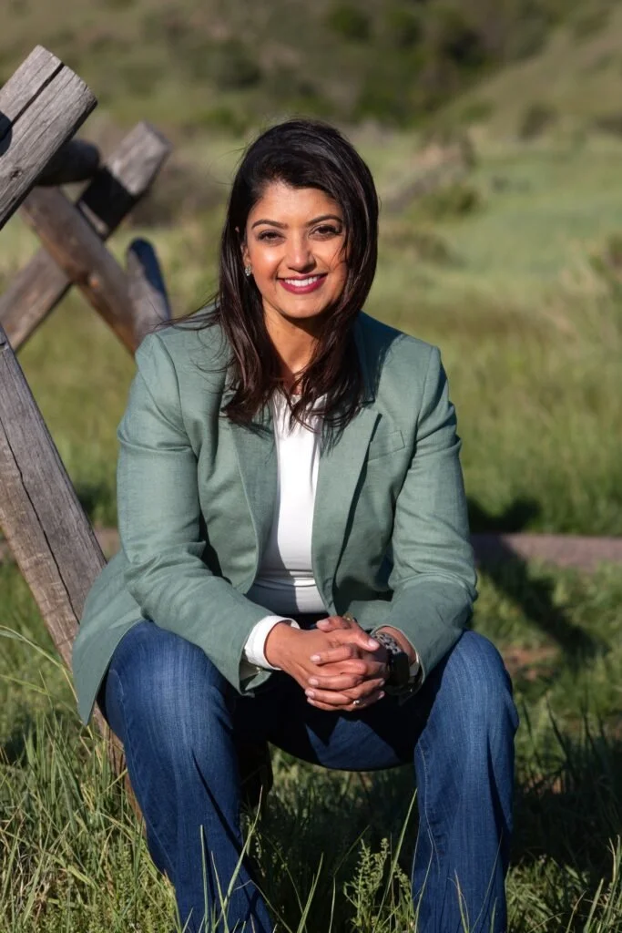 Woman in green blazer sitting outdoors on grassy field with a wooden structure in the background