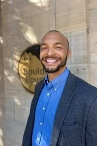 A man with a beard smiling in front of a wall with a sign that reads 'Boulder Cour'.