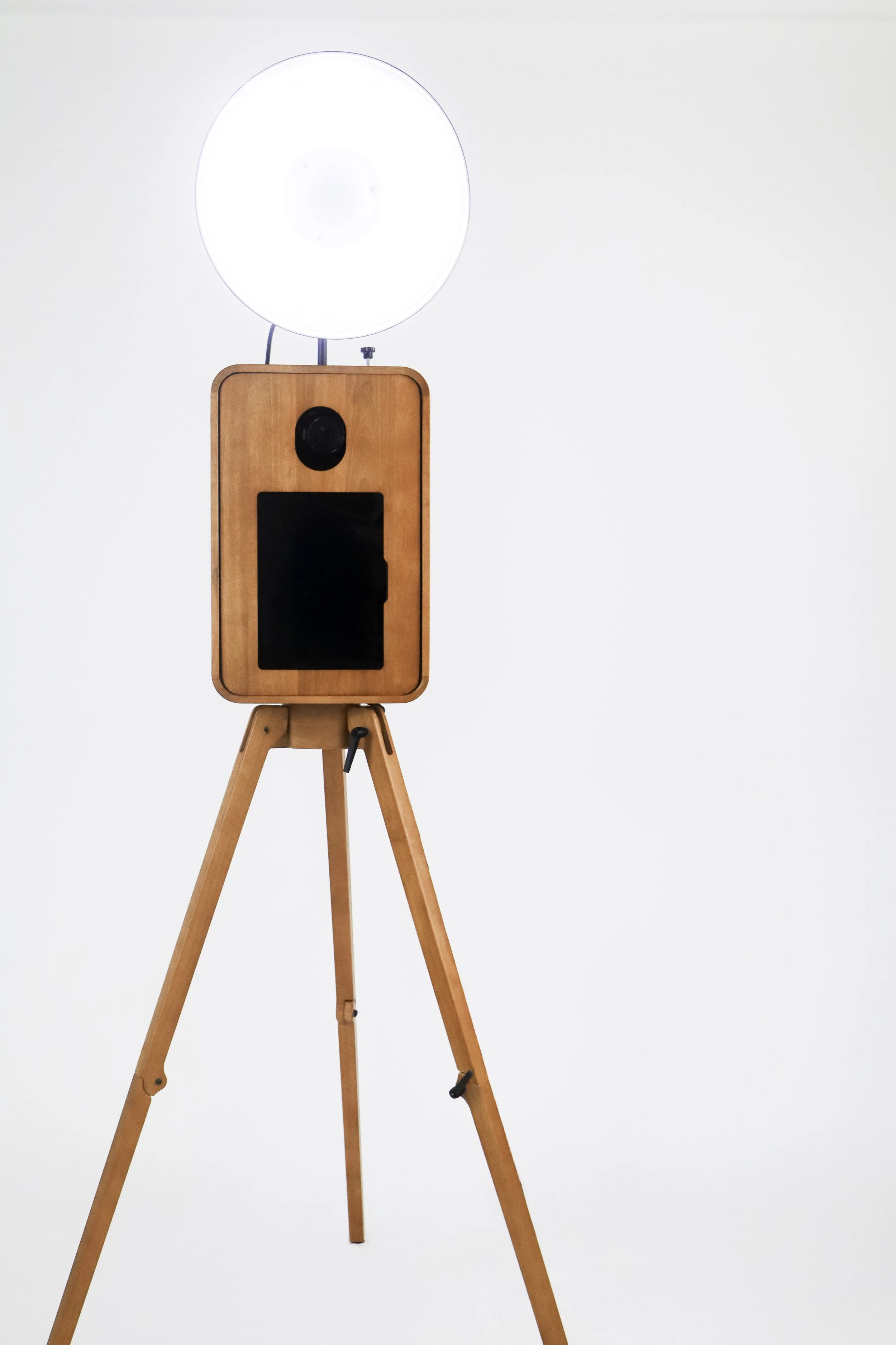 A wooden tripod with a circular white light and a black camera on top, against a plain white wall.