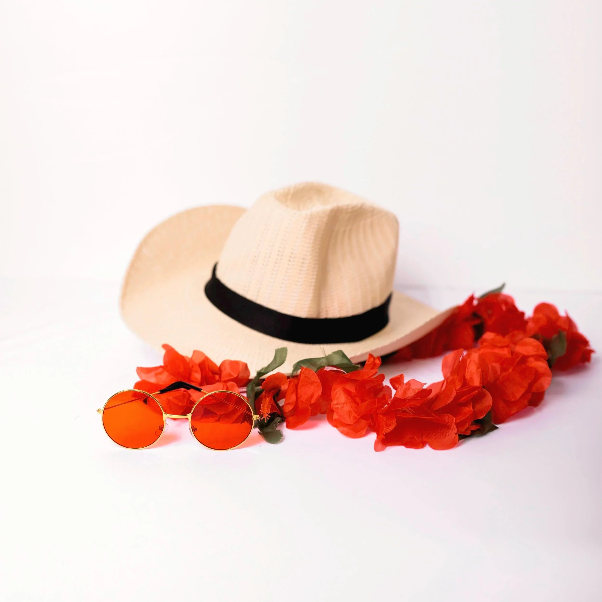 A pair of orange sunglasses, a wide-brimmed straw hat with black trim, and red flowers are arranged on a white background.