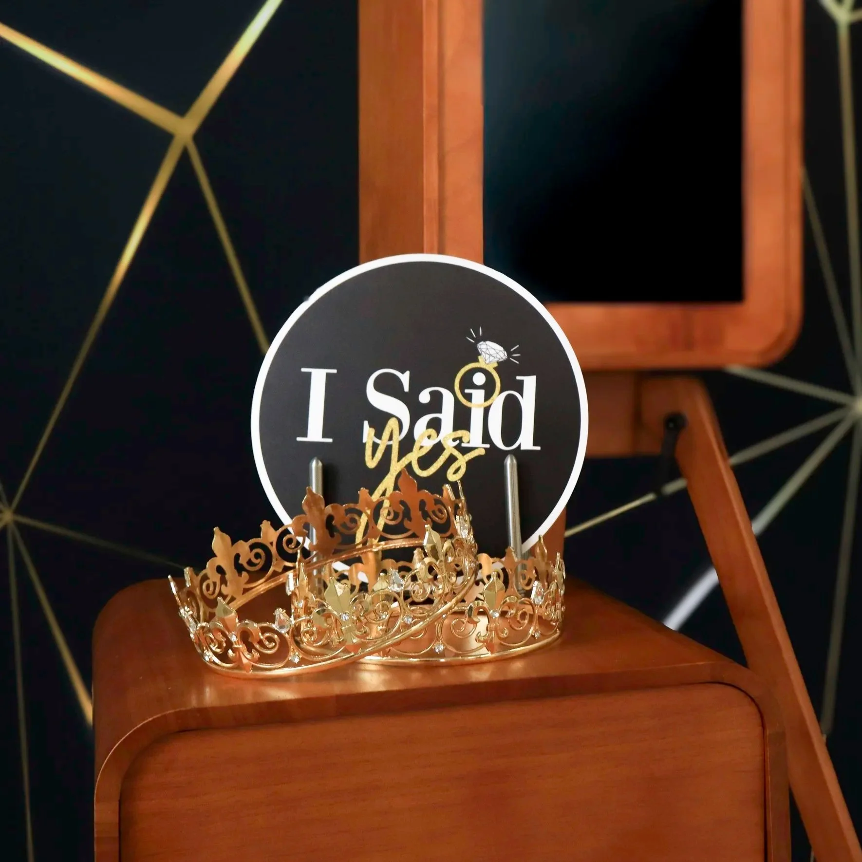 A gold crown with intricate swirling patterns and embedded crystals resting on a wooden surface. Behind the crown, there is a black and white wall clock with gold hands, a decorative design featuring a diamond and the words 'I Said Yes,' and a wooden chair with a black seat cushion.