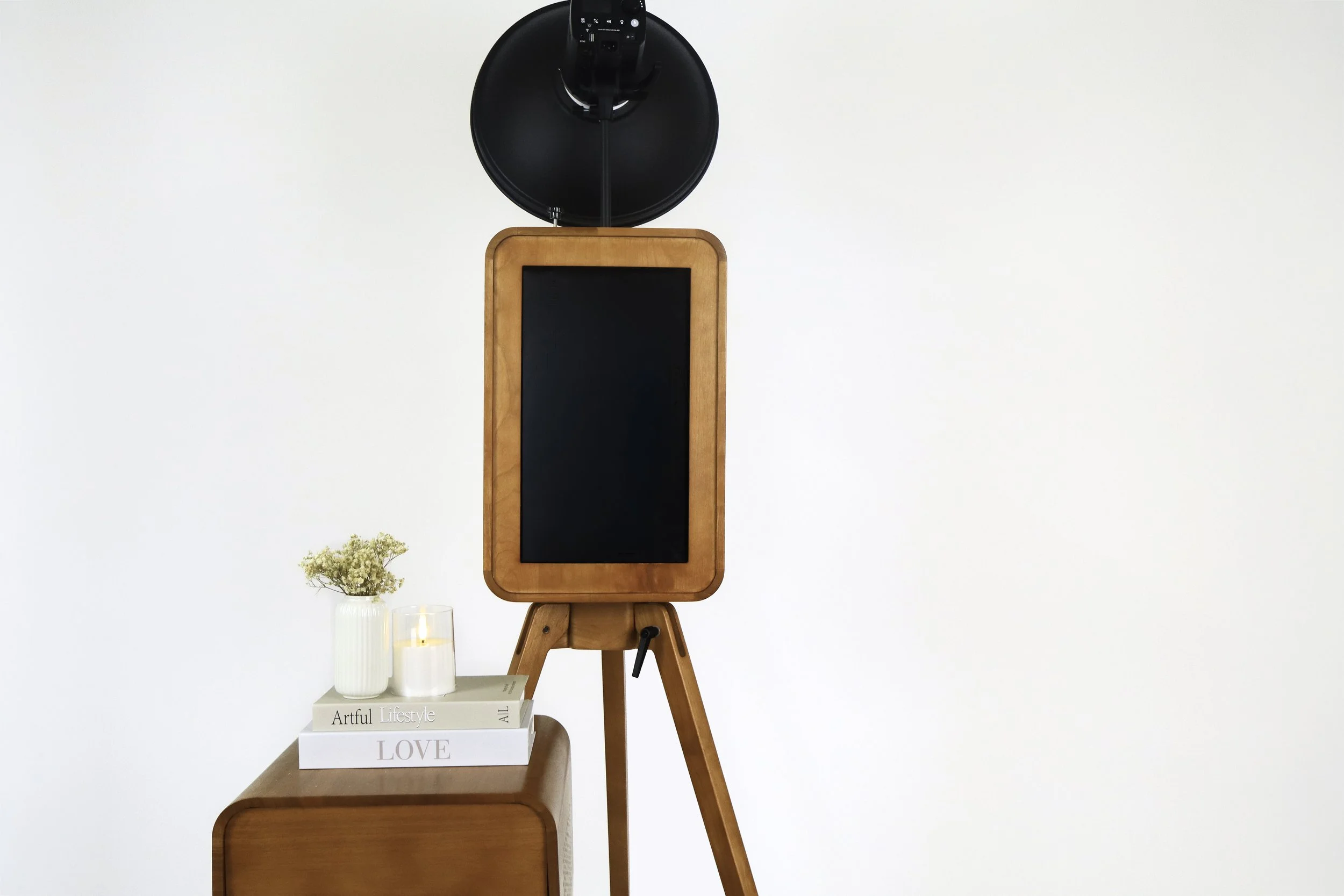 A wooden photo stand with a black chalkboard space, on a tripod, against a white wall. To the left are coasters, candles, and books with the words 'Artful Lifestyle' and 'LOVE' visible. There is a small vase with flowers.