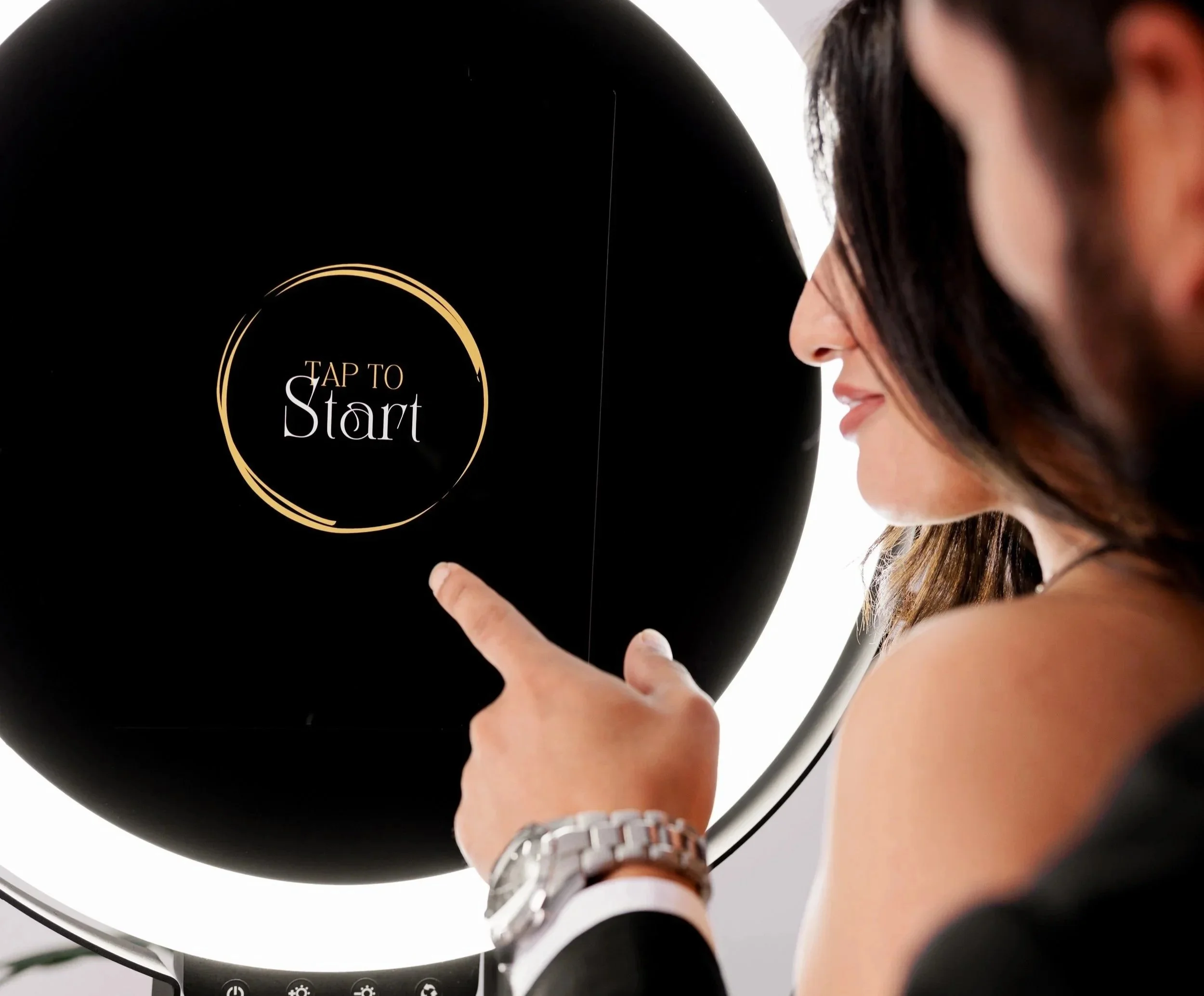 A woman with long dark hair and a watch on her wrist is pressing a button on a black object with a circular gold outline and the text 'Tap to Start.'
