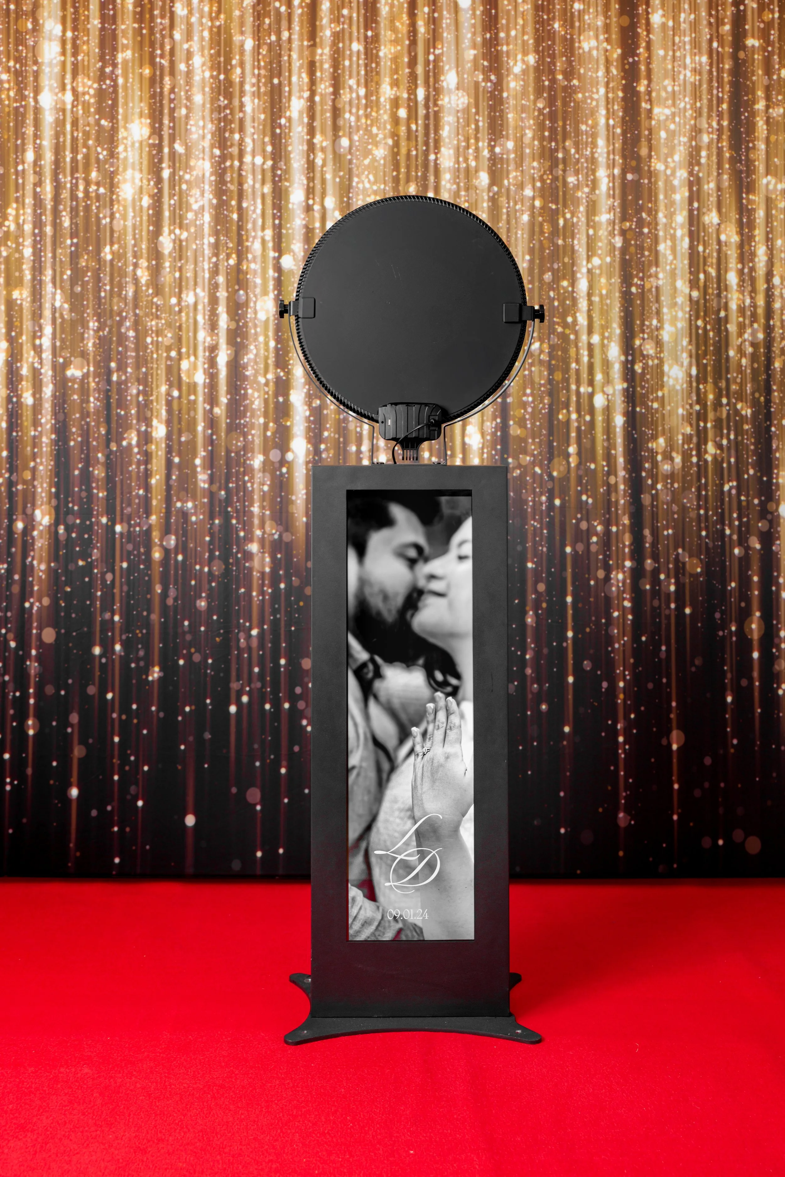 Modern Photo booth. A Toronto photo booth with a black-and-white picture of a couple about to kiss, with a date 09.01.24 at the bottom. The booth is on a red carpet with a gold glitter backdrop.