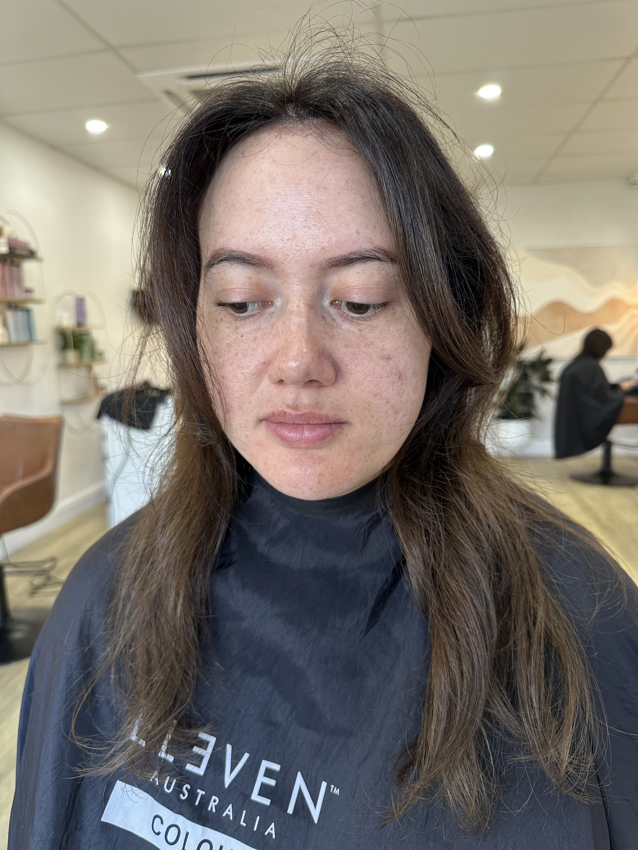 A woman with light skin and brown hair sitting in a salon chair, wearing a black cape with. This is a before hair extensions photo that shows the hair is fine
