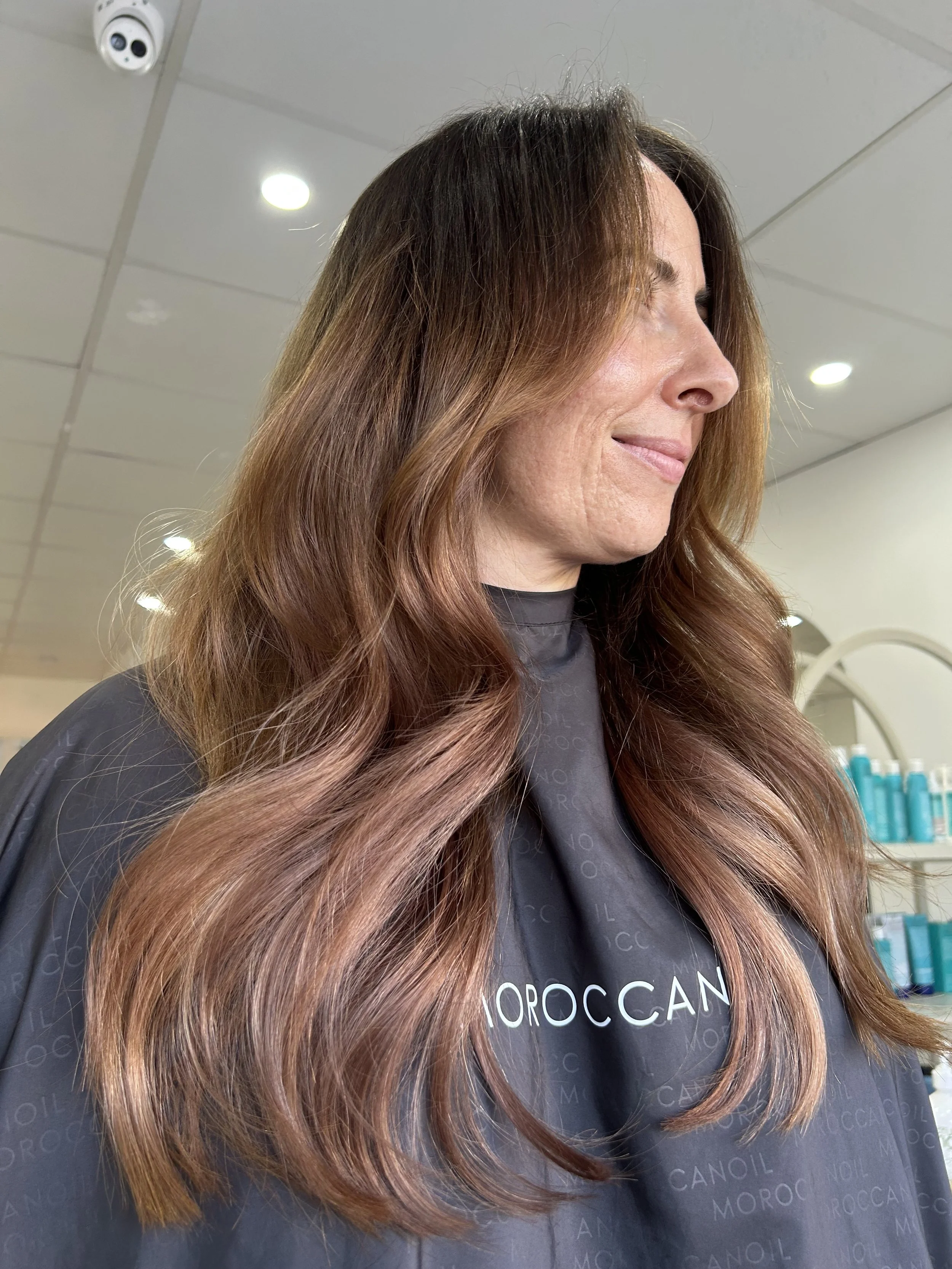This is an after photo of a client who has had hair extensions. A woman with long, wavy, auburn hair in a salon, wearing a black salon cape with the words 'Moroccan' printed on it.