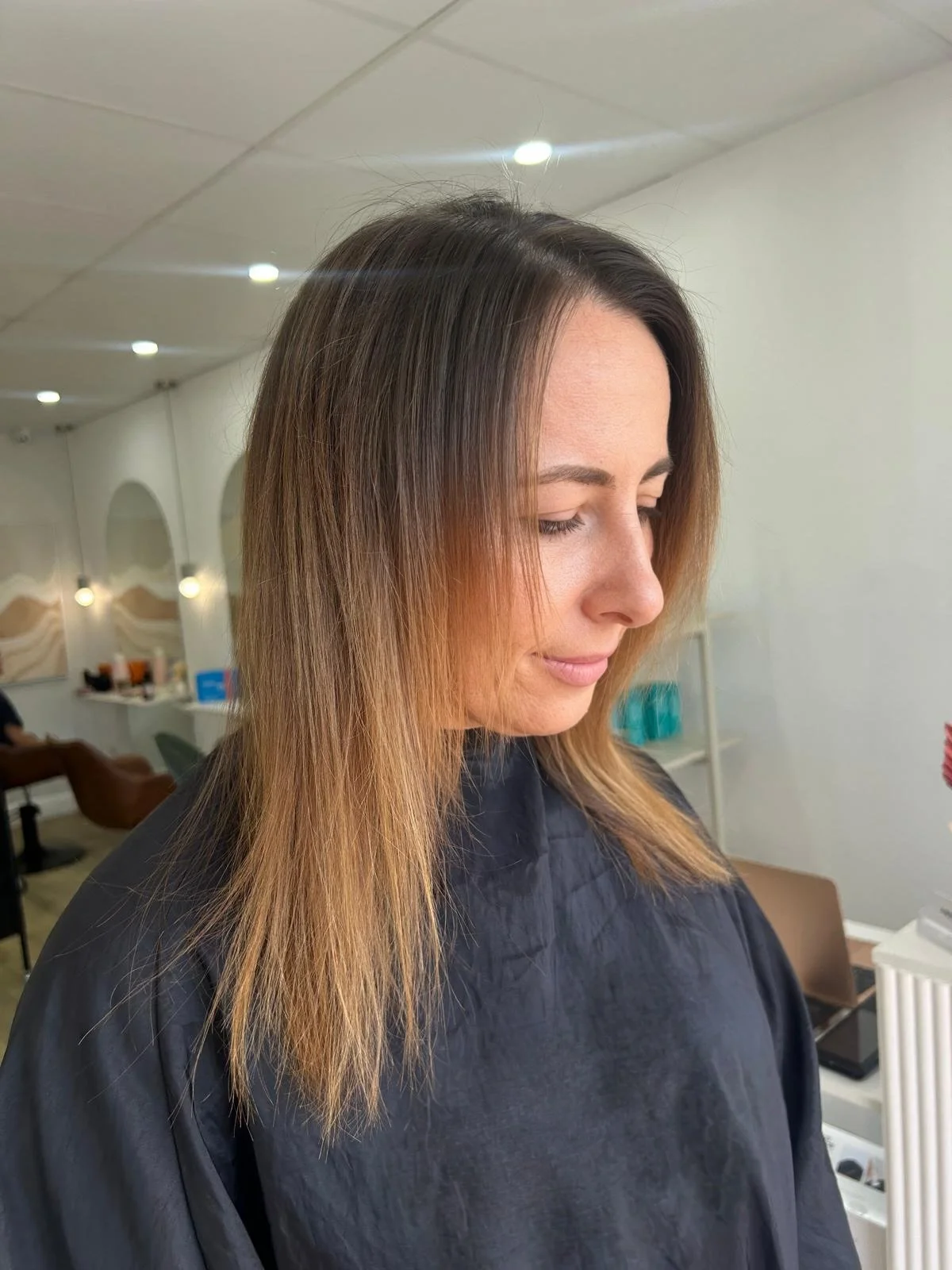 This is the before photo of a client who is getting extensions. Her hair is fine and whispy on the ends. A woman with shoulder-length brown hair with blonde highlights is sitting in a salon chair, wearing a black salon cape, with her eyes closed.