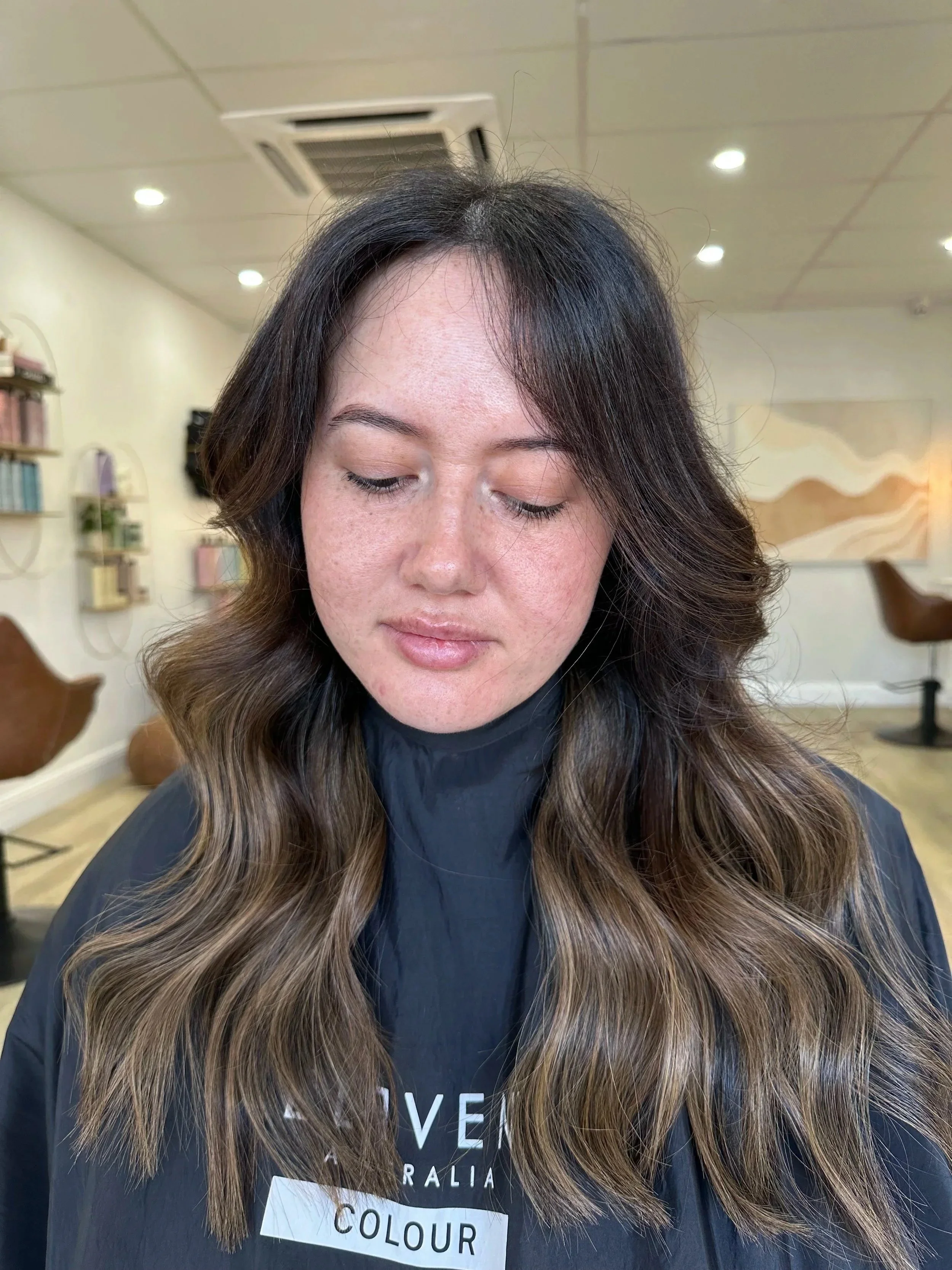 This is the after photo from an extensions make over. A woman with long wavy brown hair with blonde highlights and closed eyes, sitting in a hair salon.
