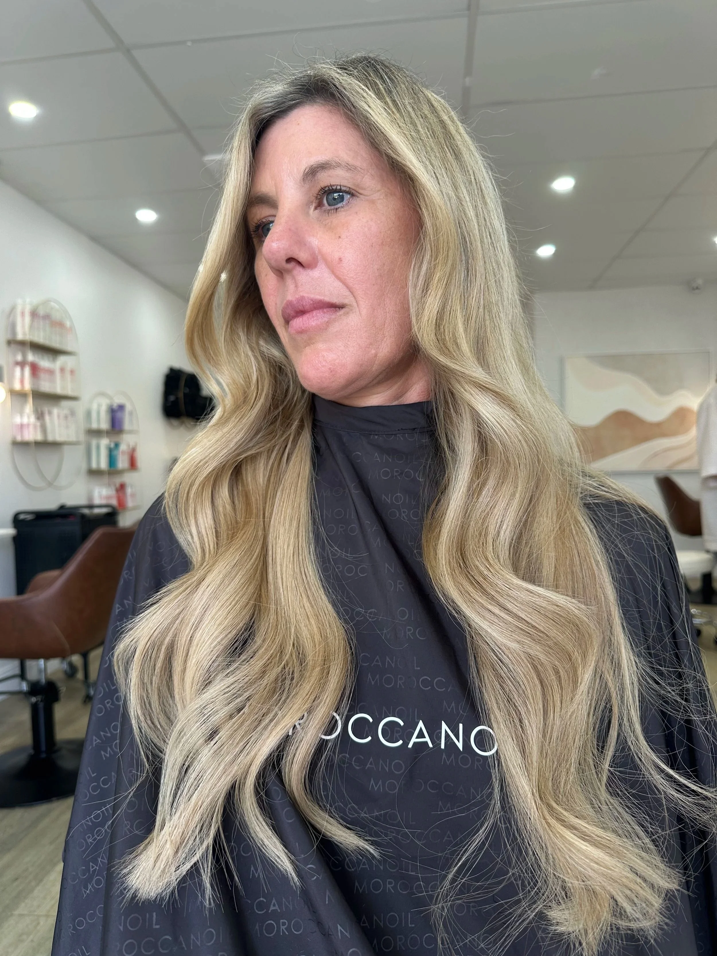 This is the same client as before only now she has extensions for lenght .Woman with long, wavy blonde hair sitting in a salon chair, wearing a black salon cape with 'MOCCANI' printed on it, in a hair salon