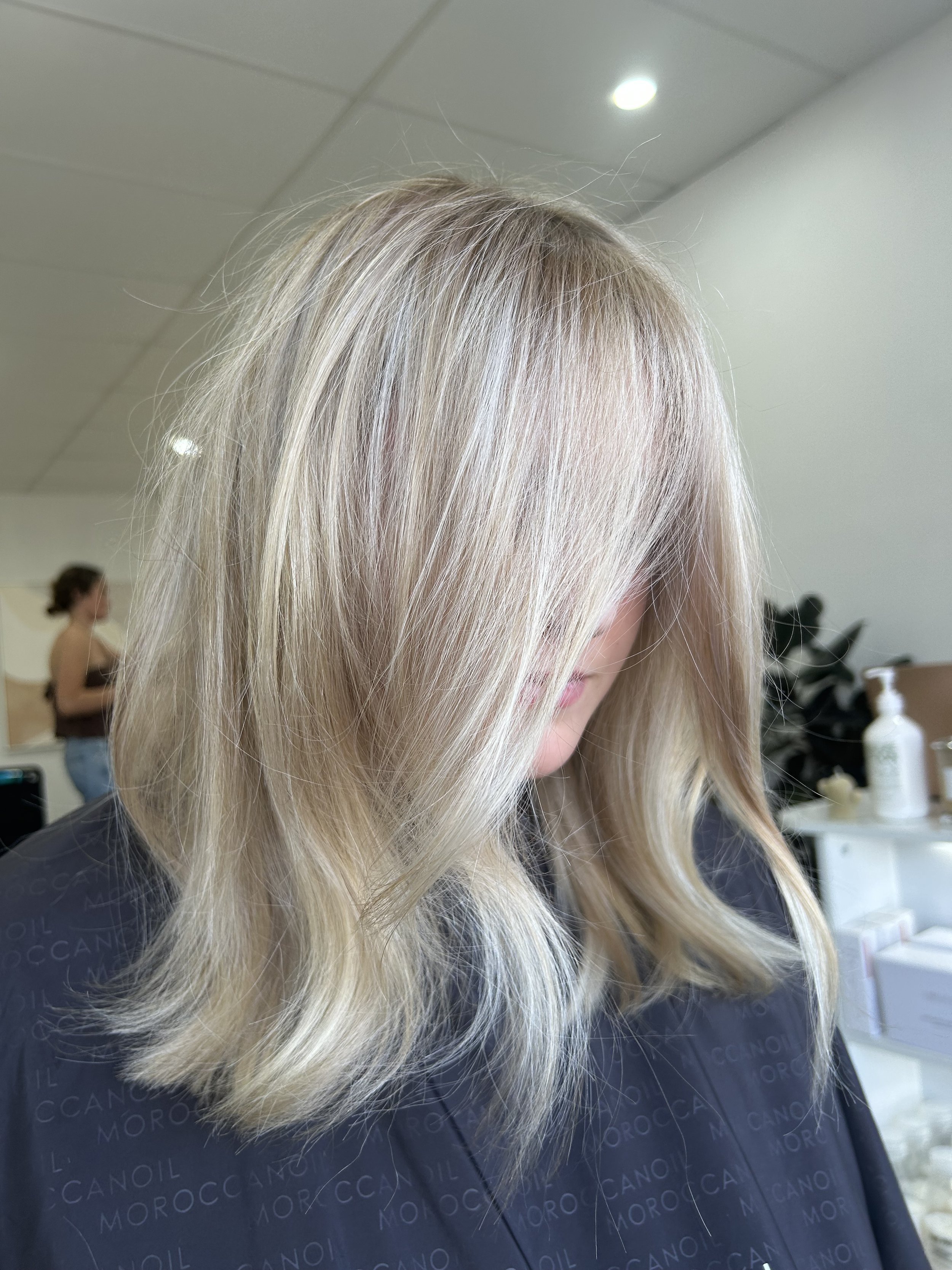 Woman with shoulder-length blonde hair in salon, with another person working in the background.