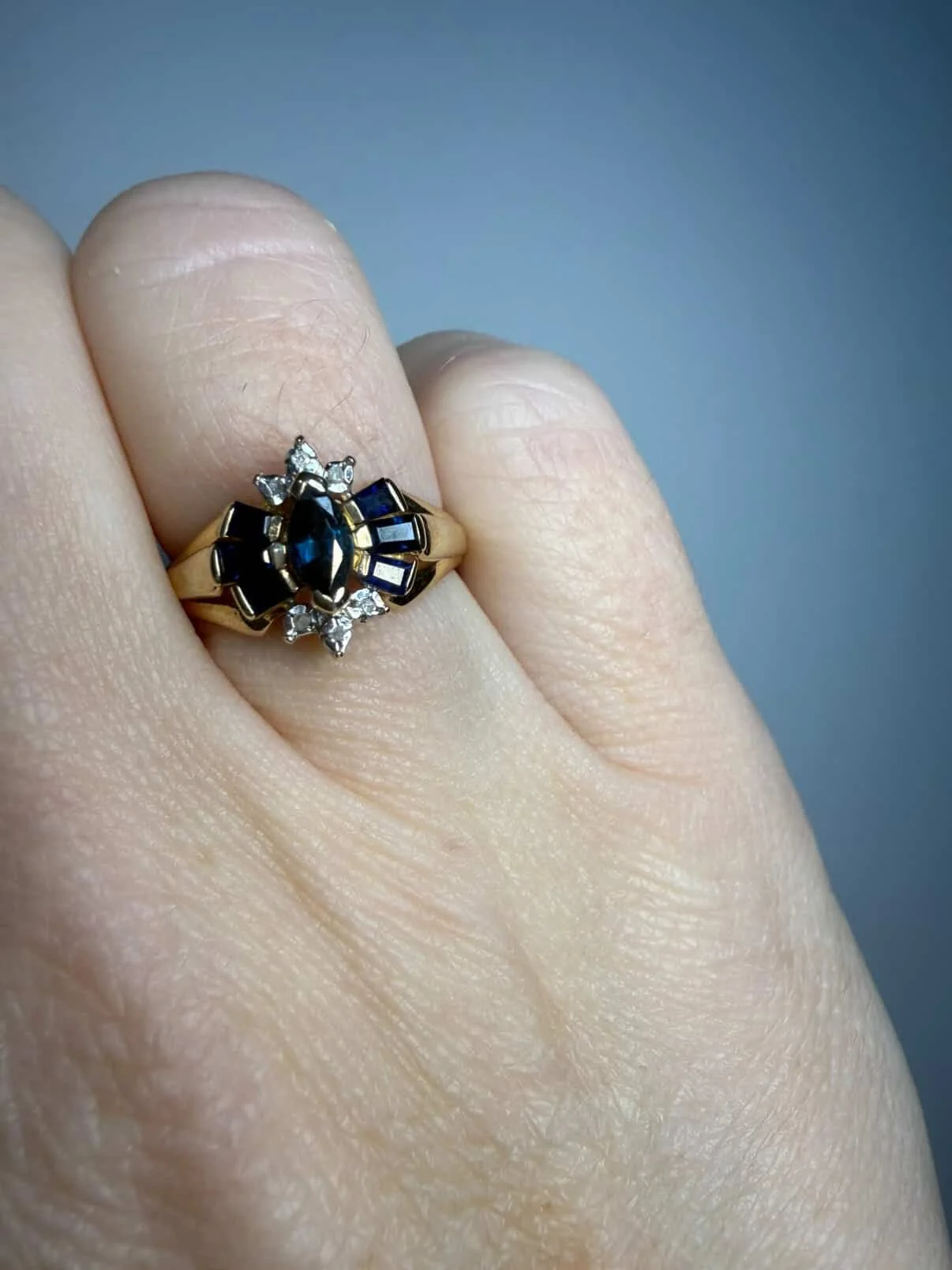A person wearing a gold, sapphire, and diamond ring on their ring finger.