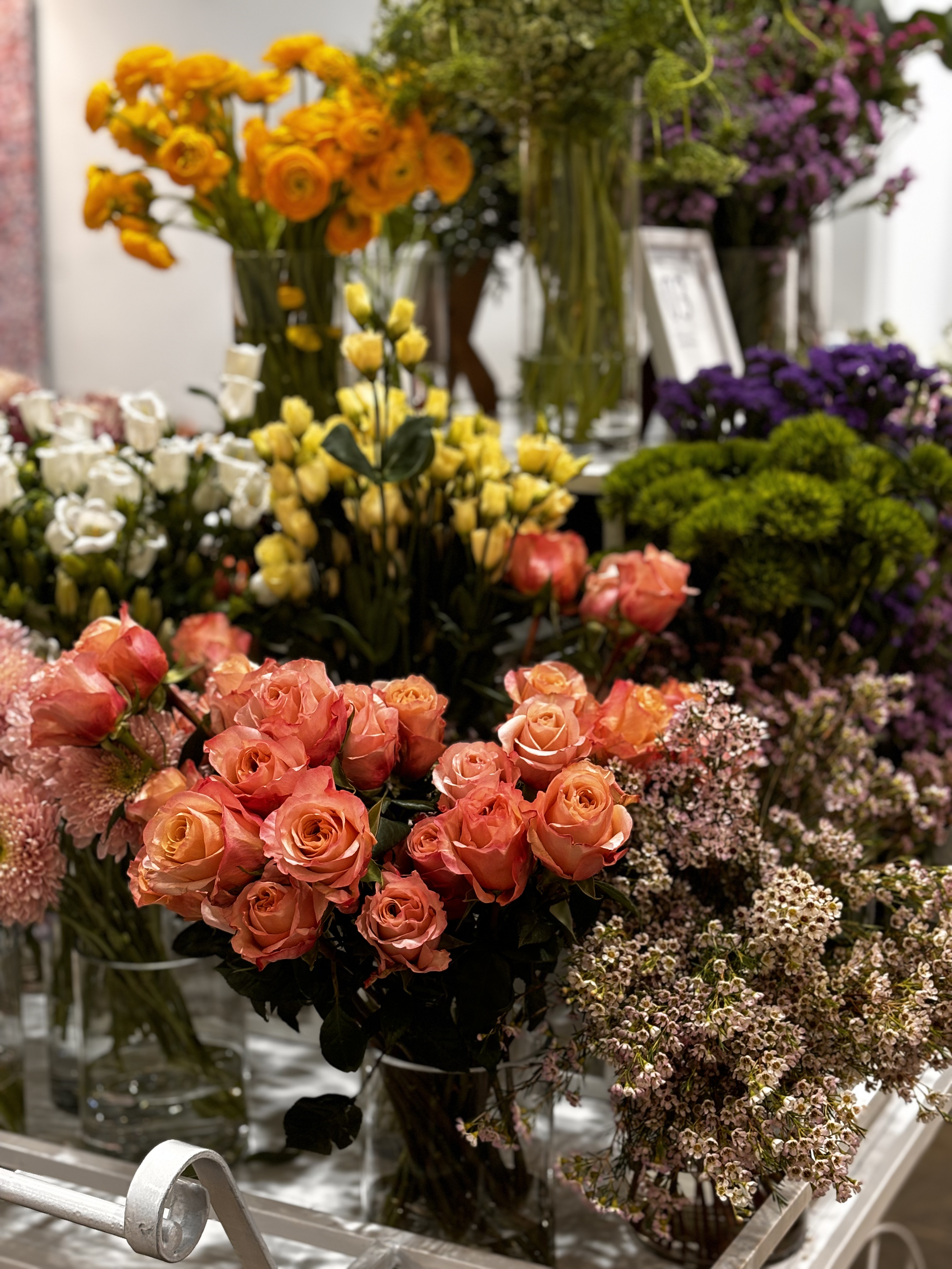 Colorful display of various fresh flowers in vases, including pink roses, white and yellow tulips, purple, white, and pink blossoms, and green foliage, arranged on a table at a flower shop.