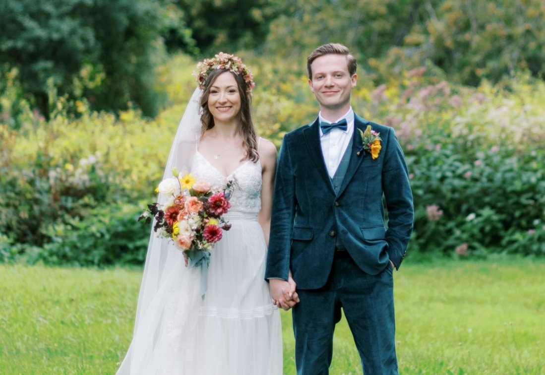 A bride and groom holding hands outdoors surrounded by greenery and flowers, smiling at the camera.