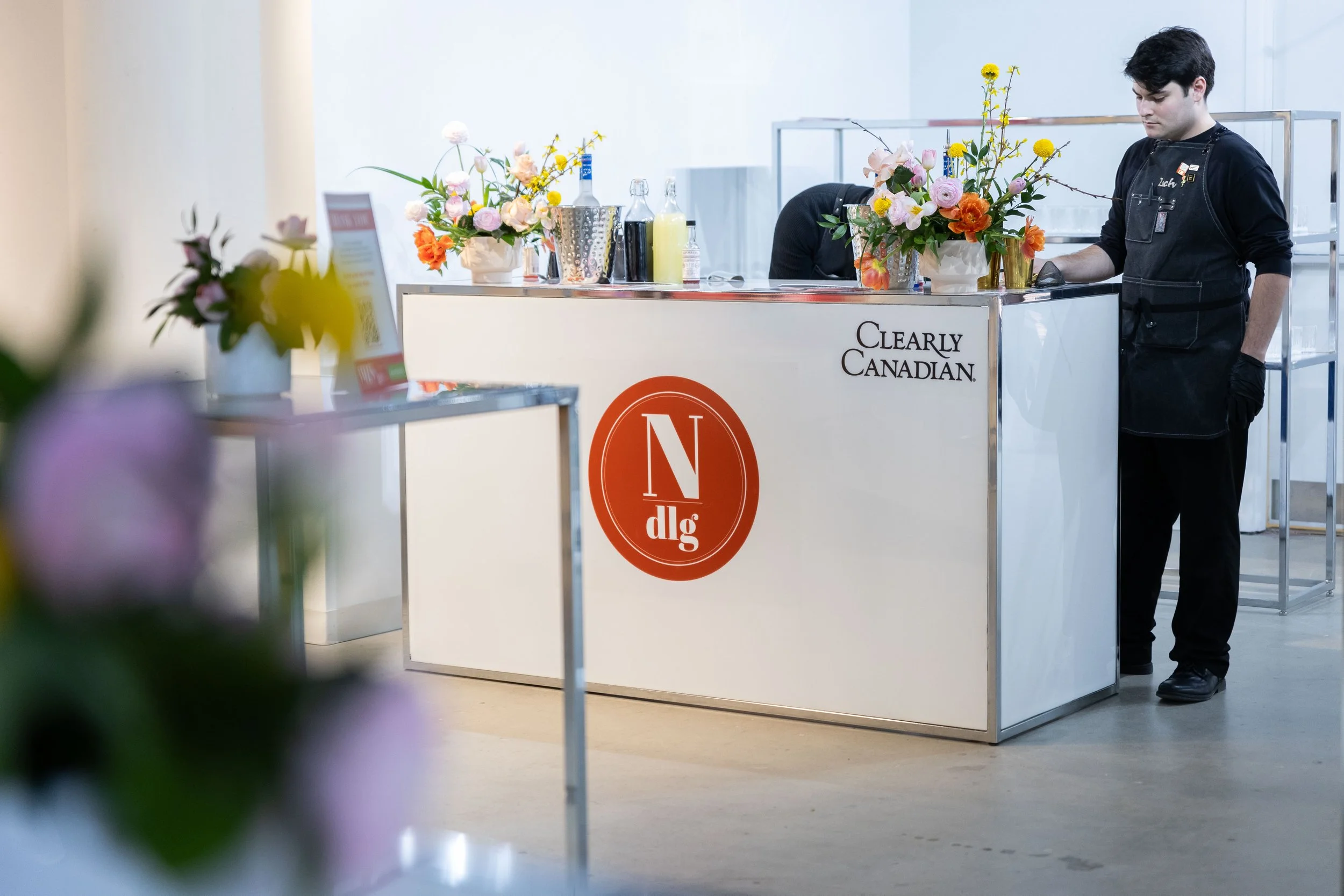 A woman at a floral display booth with a man working behind the counter, decorated with colorful flowers and bottles, at an event or market.