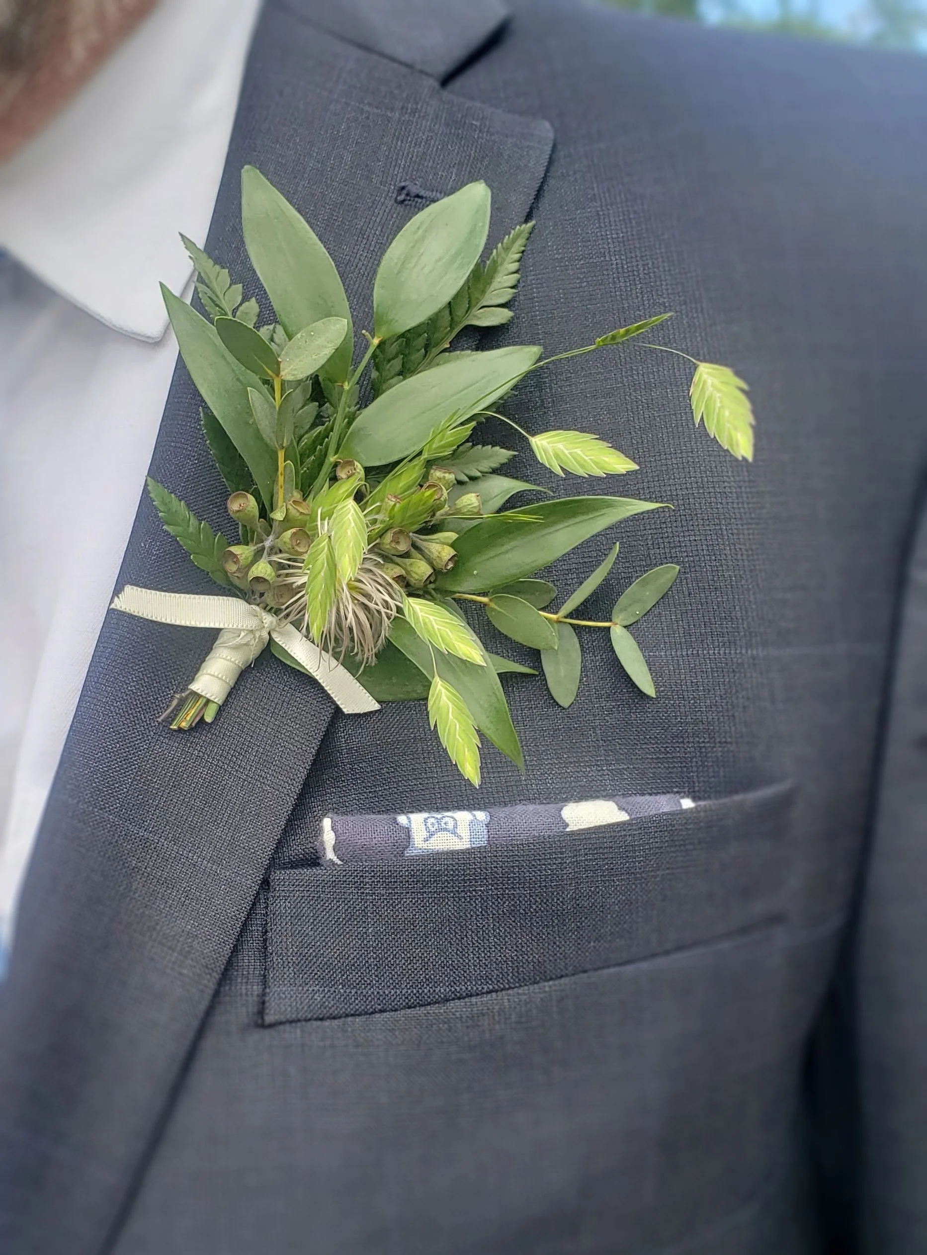 A close-up of a dark suit jacket with a boutonniere made of clematis buds and wood oat grass pinned to the lapel. 