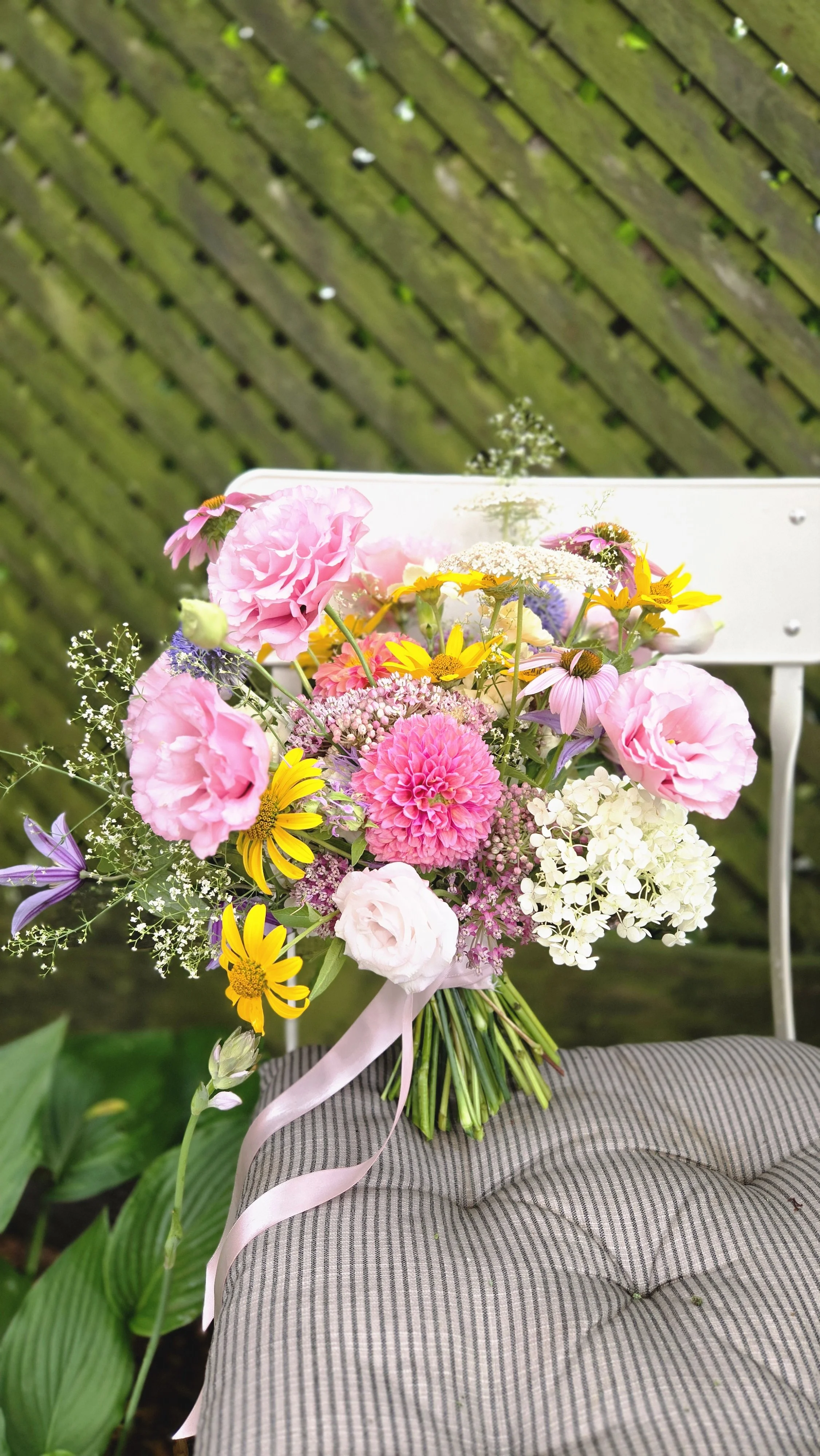 A bouquet of pink, yellow, white, and purple flowers resting on a striped gray cushion outdoors, with a mossy wooden fence in the background.