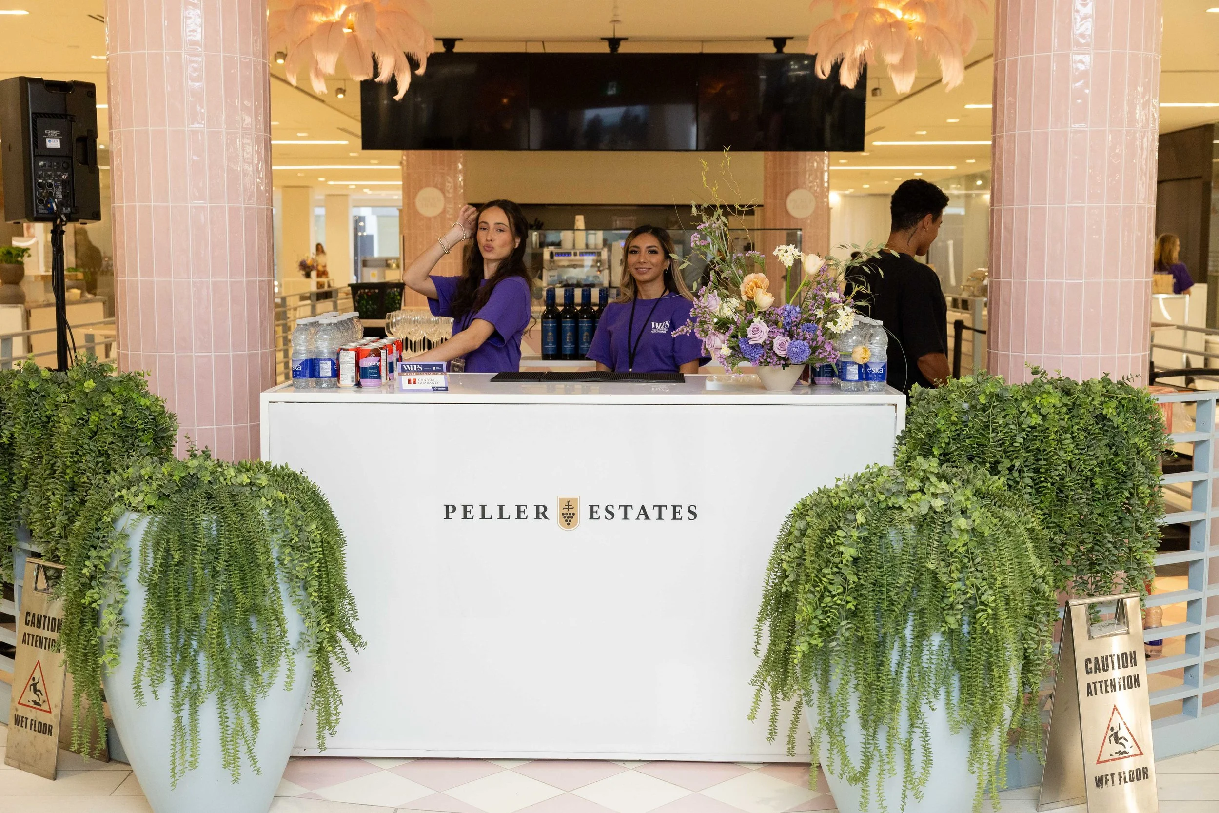 A reception desk for Peller Estates with two women in purple shirts standing behind it, one making a playful face and the other smiling. The desk is decorated with two large green fern plants in white pots and a colorful flower arrangement. There are