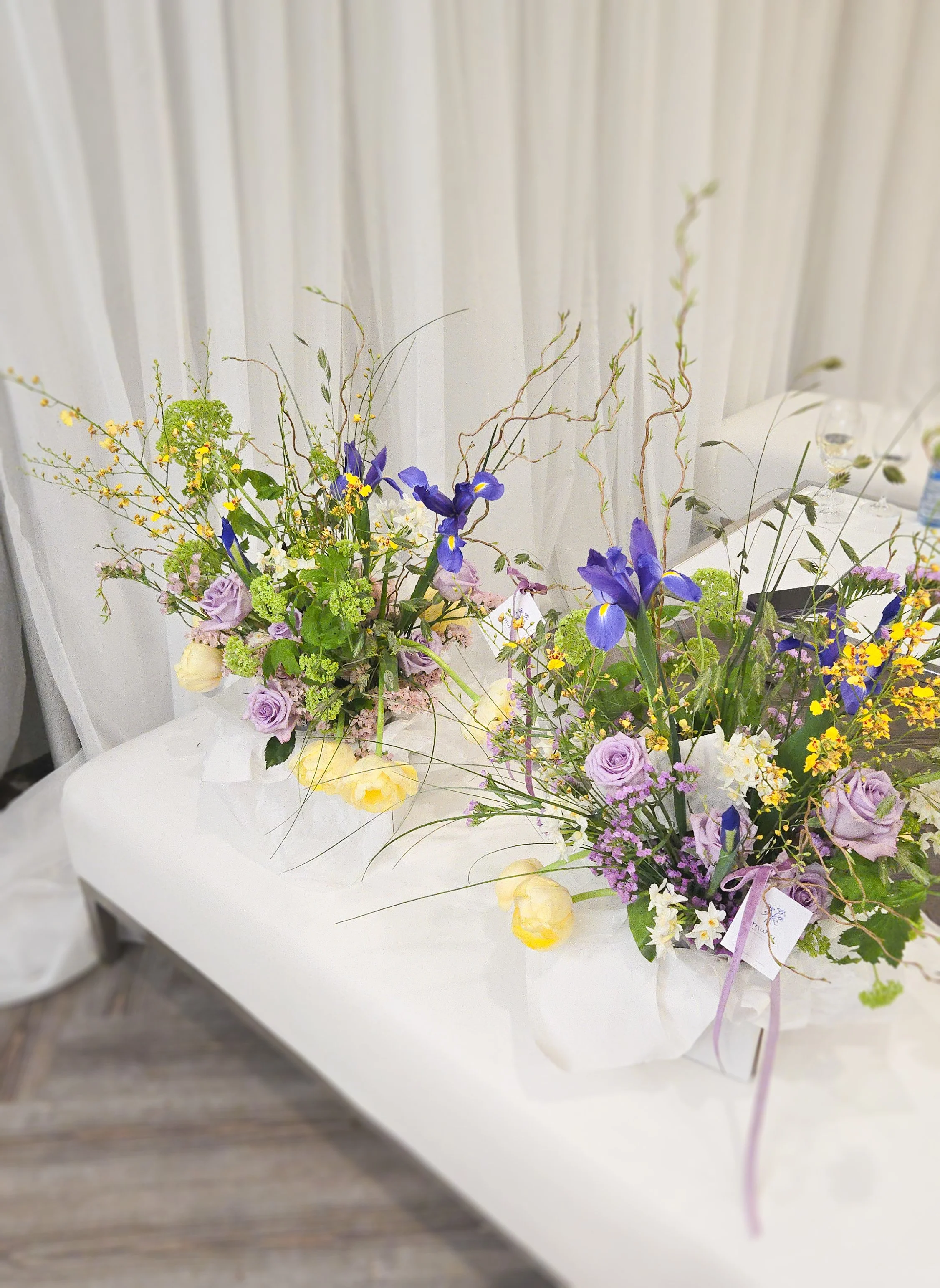 Two floral arrangements with purple roses, yellow flowers, and greenery on a white table.