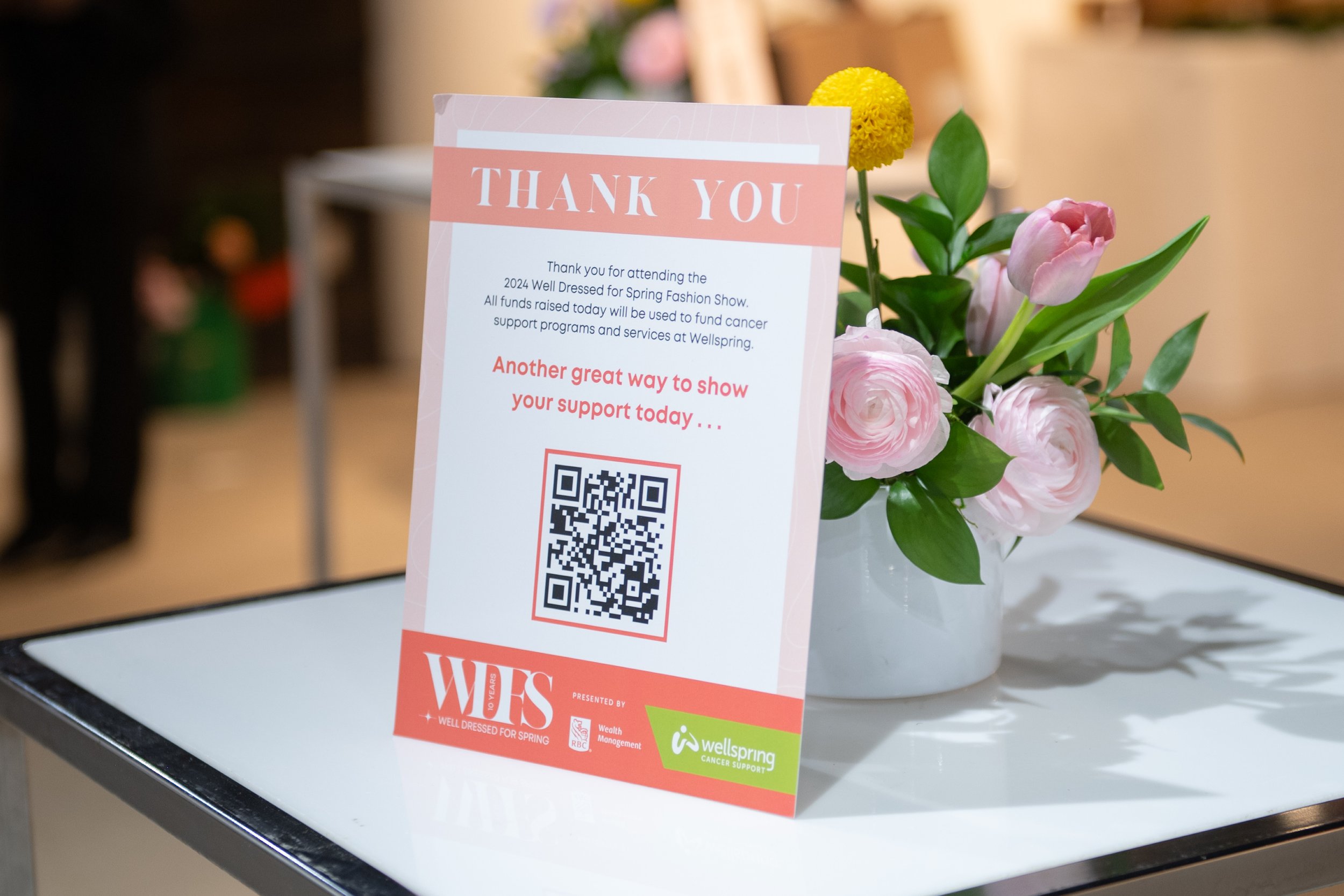 A thank you sign with a QR code placed on a white table next to a white vase with pink and yellow flowers.