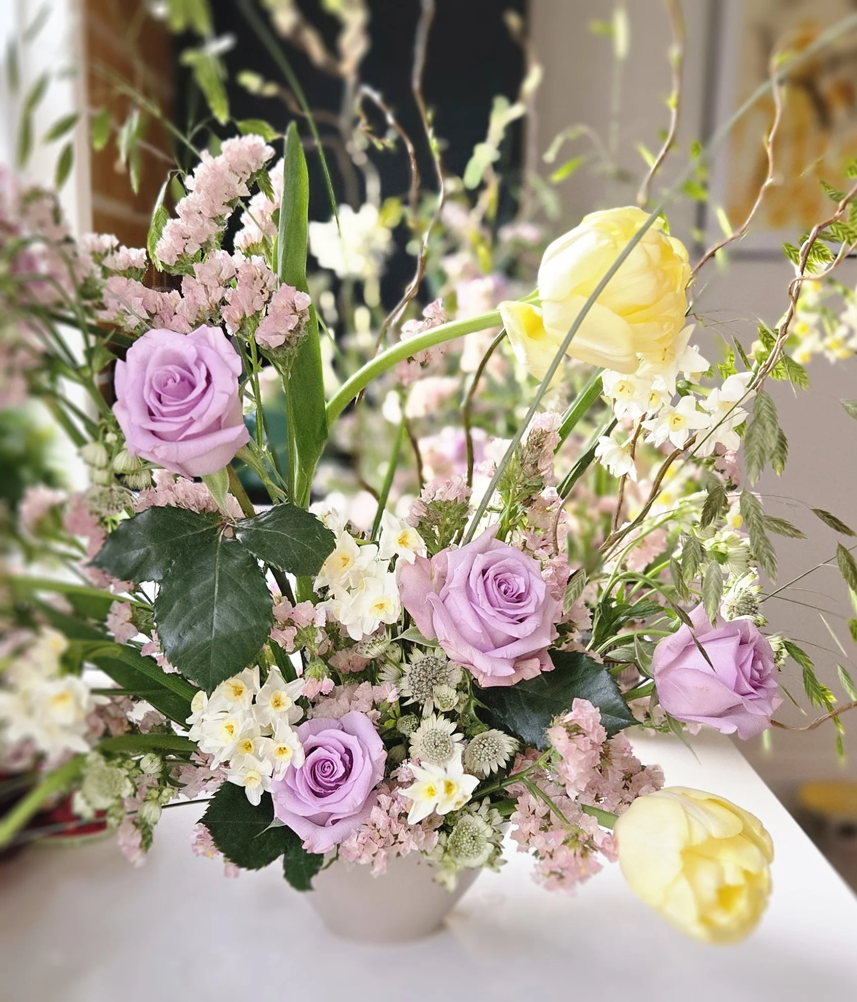 A floral arrangement with lavender roses, yellow tulips, pink and white flowers, and greenery in a white vase.