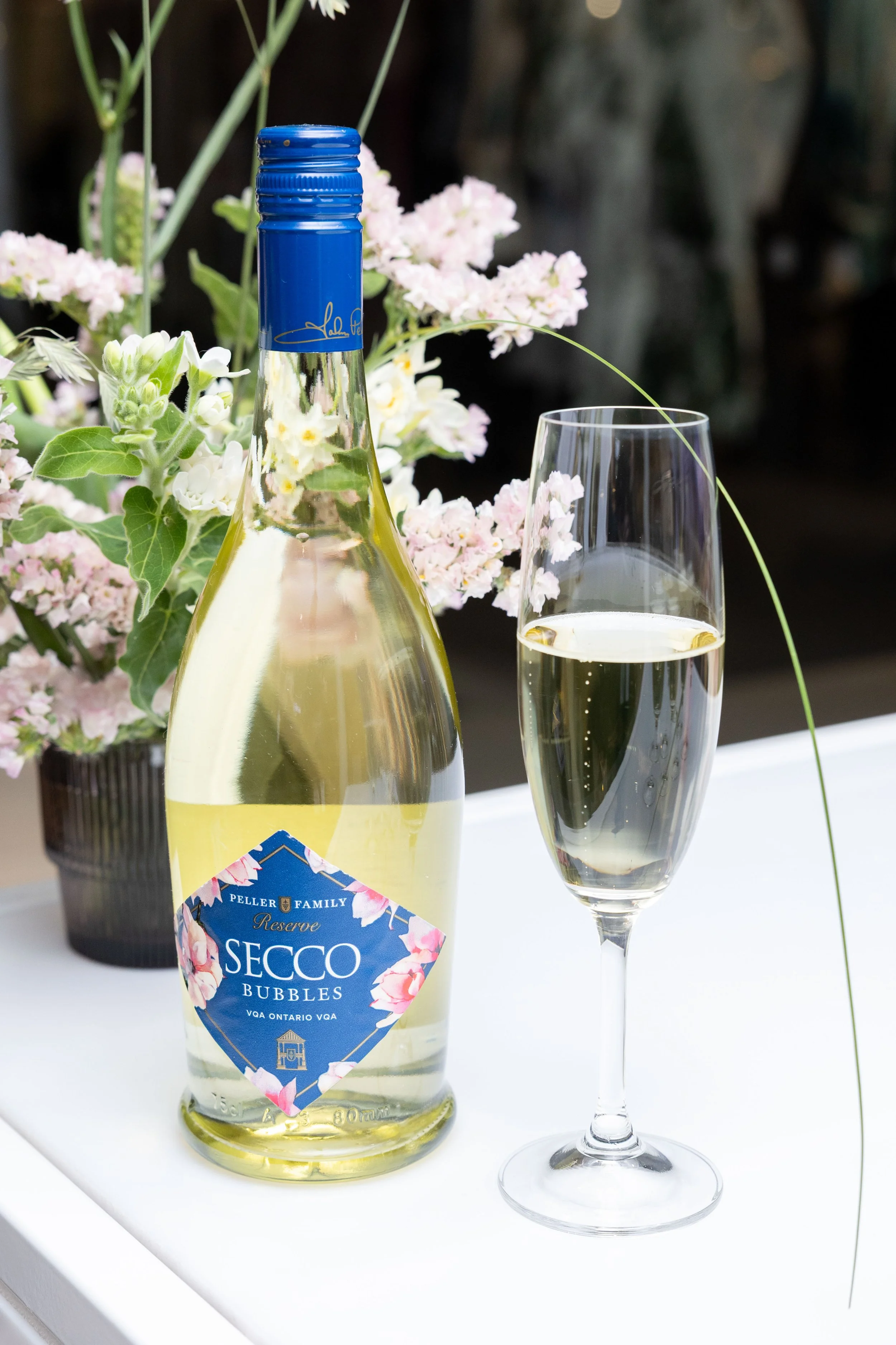 A bottle of Secco bubbles wine and a filled champagne flute on a white table with pink and white flowers in the background.