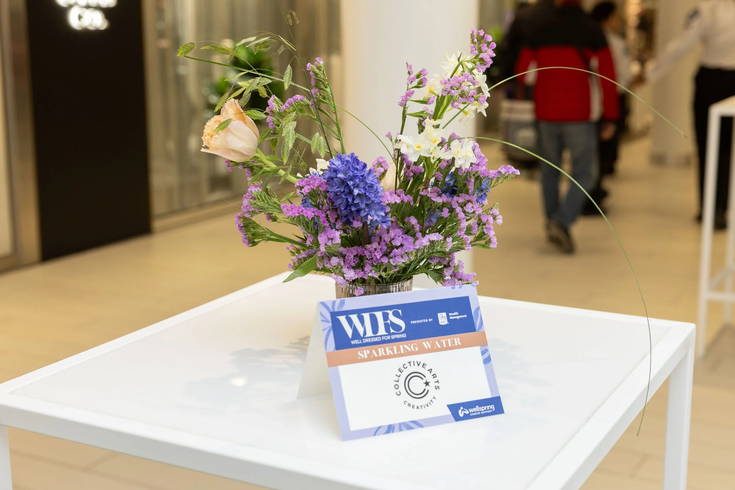 A floral arrangement with purple, blue, white, and pale pink flowers on a white table, with a sign that reads 'WFS Well Dressed for Spring Sparkling Water' and 'Collective Arts Creativity' in an indoor setting with people in the background.
