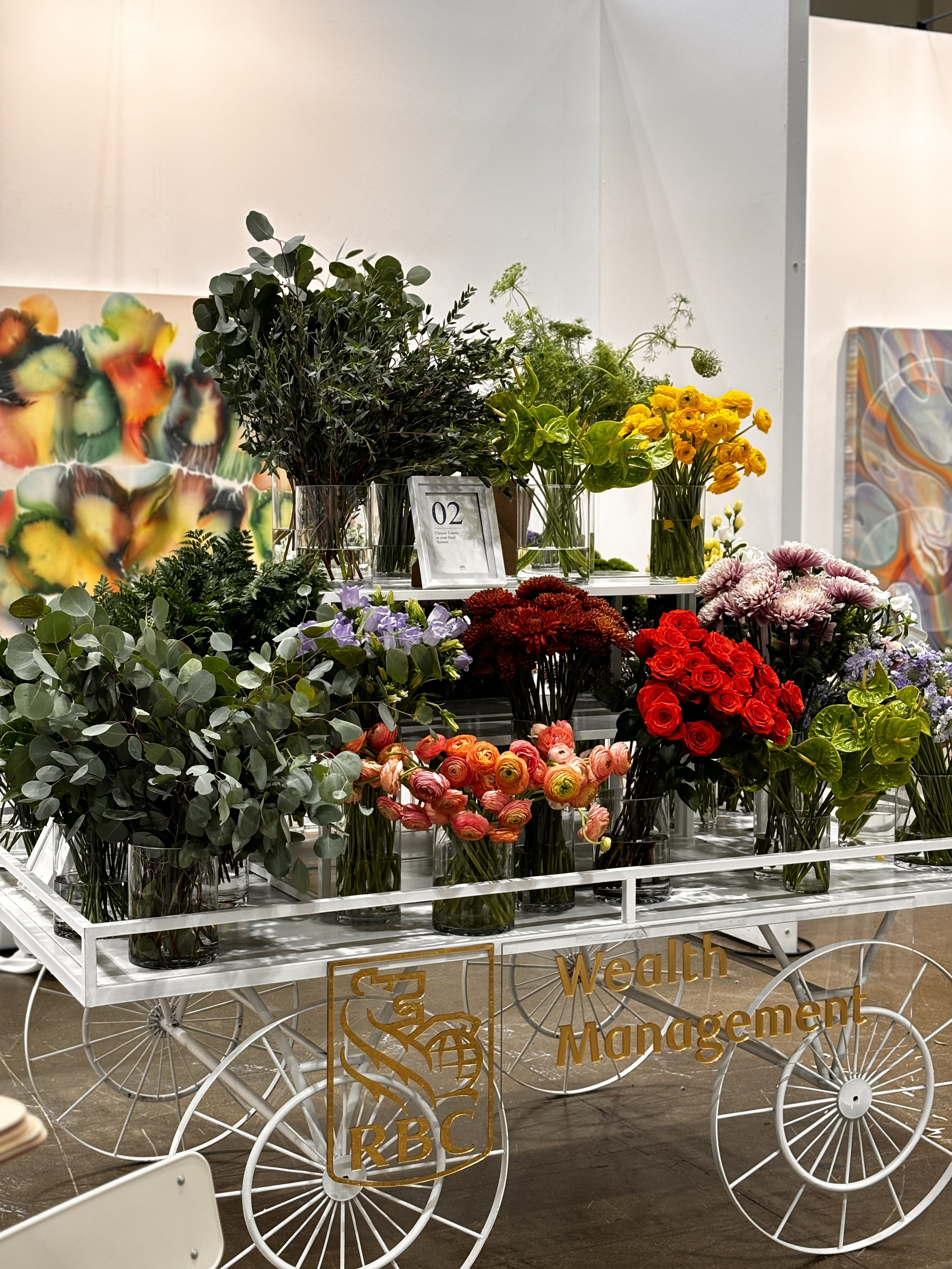 A white metal floral cart filled with various colorful flower arrangements. The cart has gold lettering that reads 'Wealth Management' and an 'RBC' logo. Behind the cart is a plain wall with some abstract artwork.