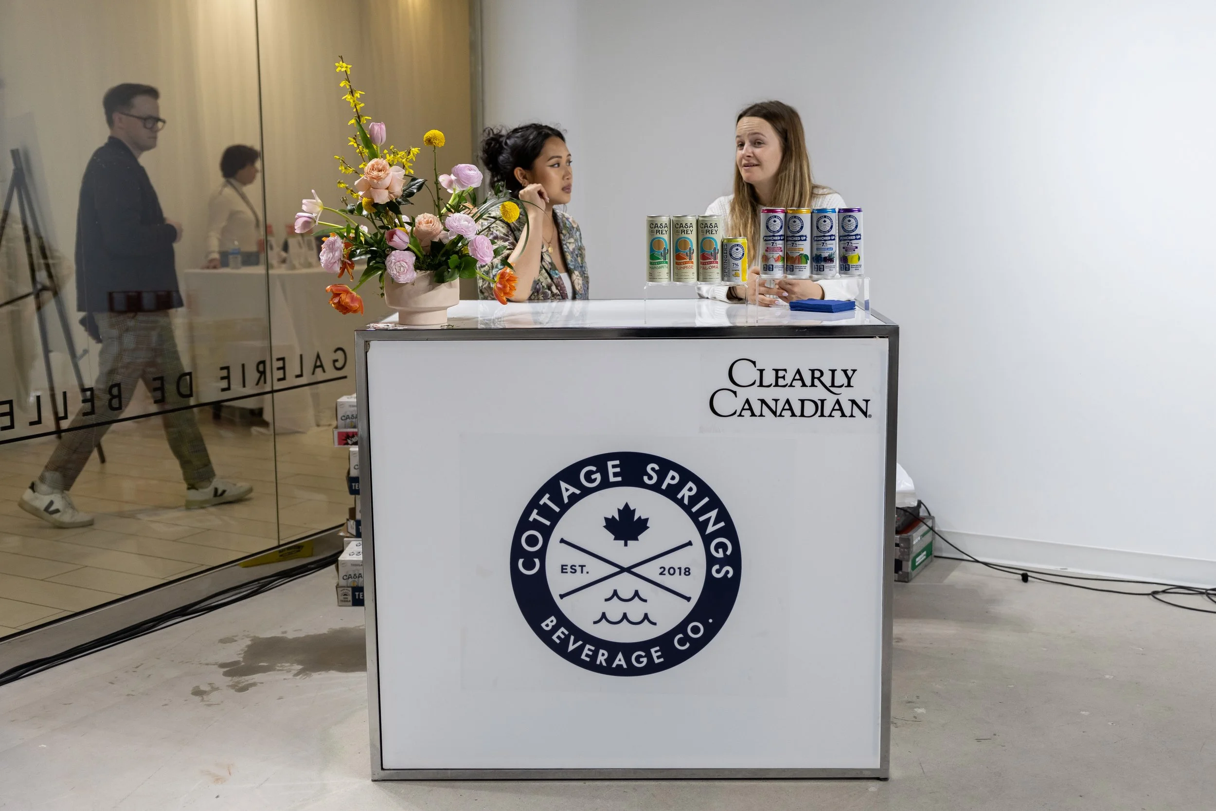 A cannabis beverage tasting booth for Cottag Springs Beverage Co. with two women speaking at the counter, a vase of colorful flowers, and canned drinks on display. The booth has a white front with logos and branding for Clearly Canadian and Cottage S