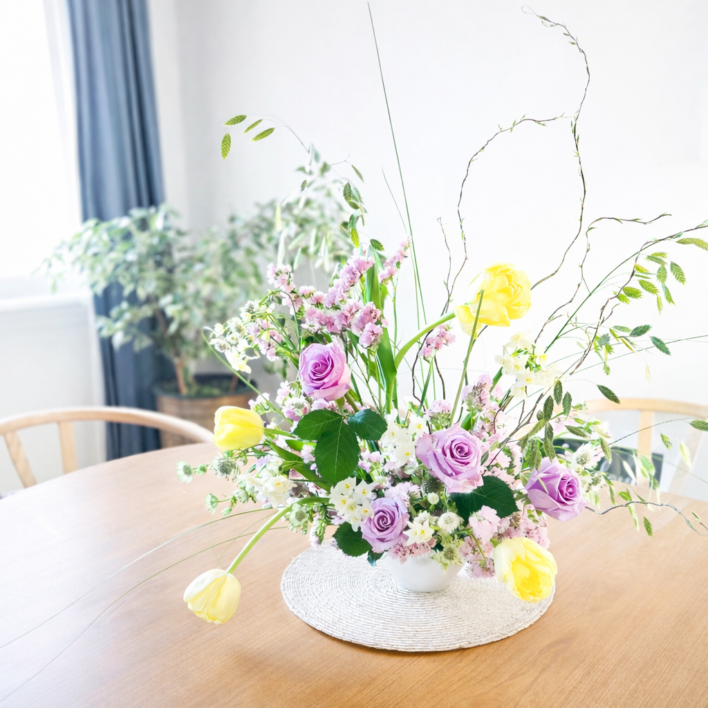 A colorful floral arrangement with purple roses, yellow tulips, white flowers, and various greenery, placed in a white vase on a white woven mat on a wooden table.