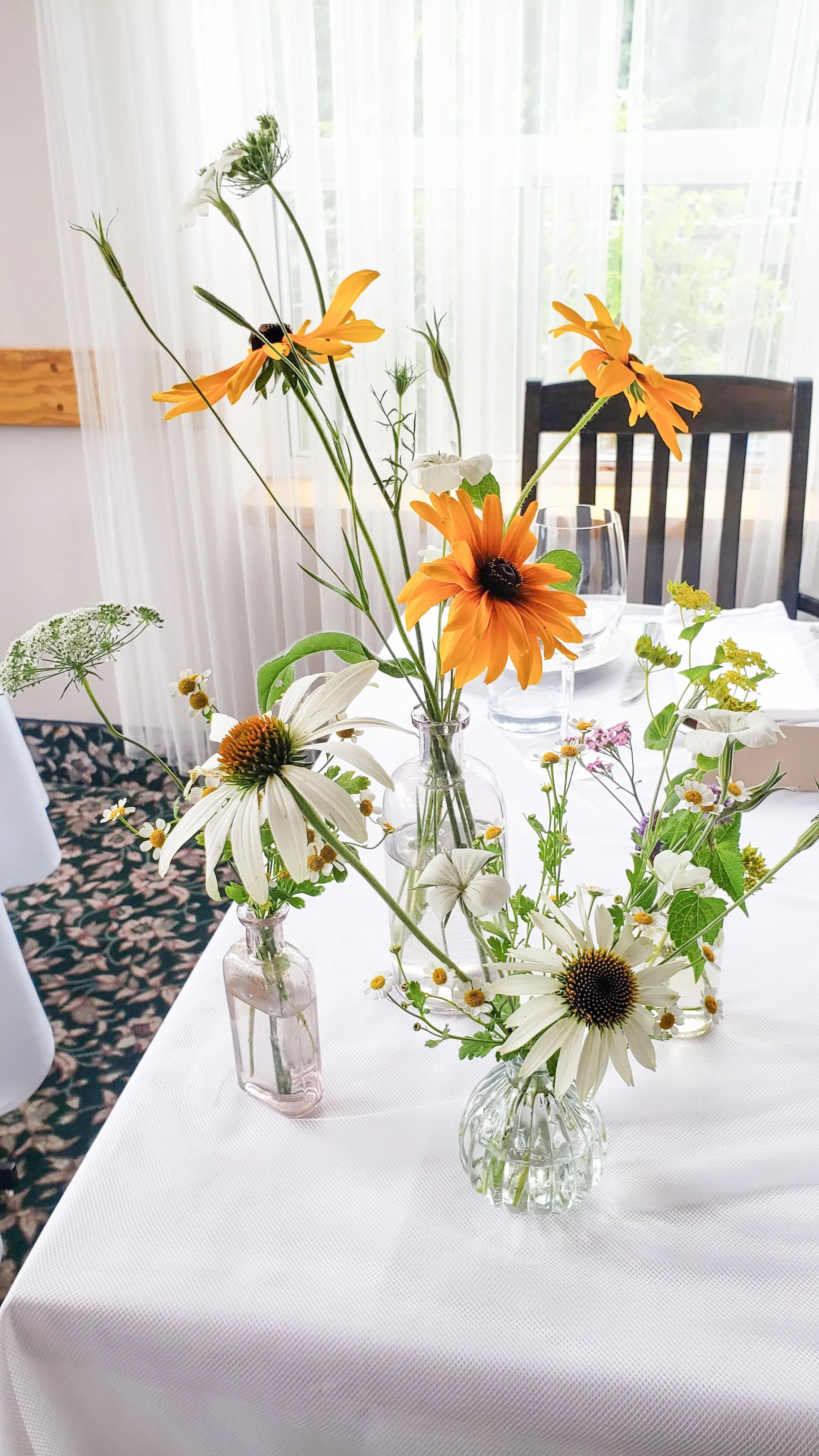 A bud vase trio with white cornflowers and yellow rudbeckia in small glass vases on a white tablecloth at a wedding.
