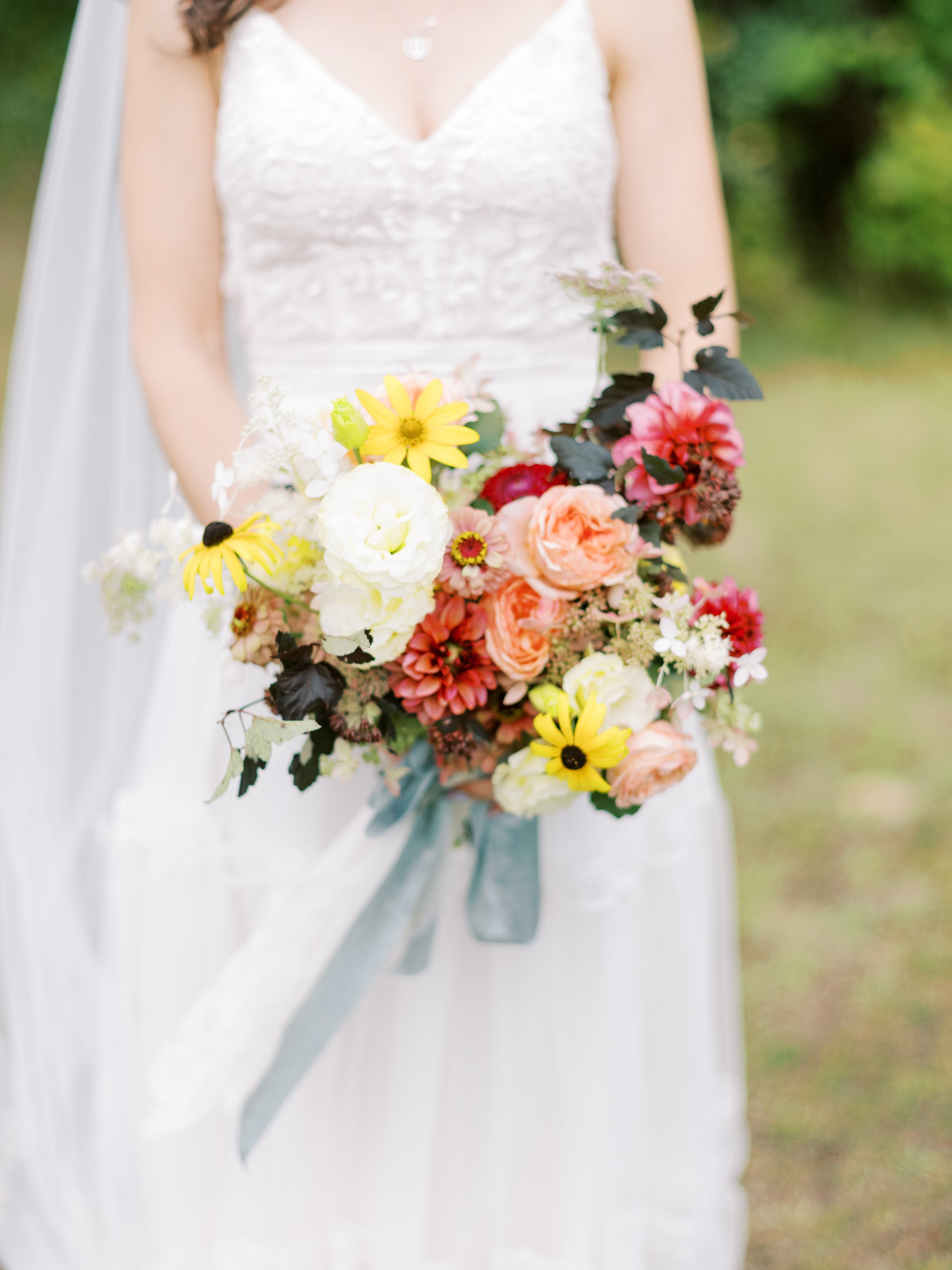 Bride in white wedding dress holding a colorful bouquet of flowers, including yellow, pink, and white blooms, tied with a blue ribbon.